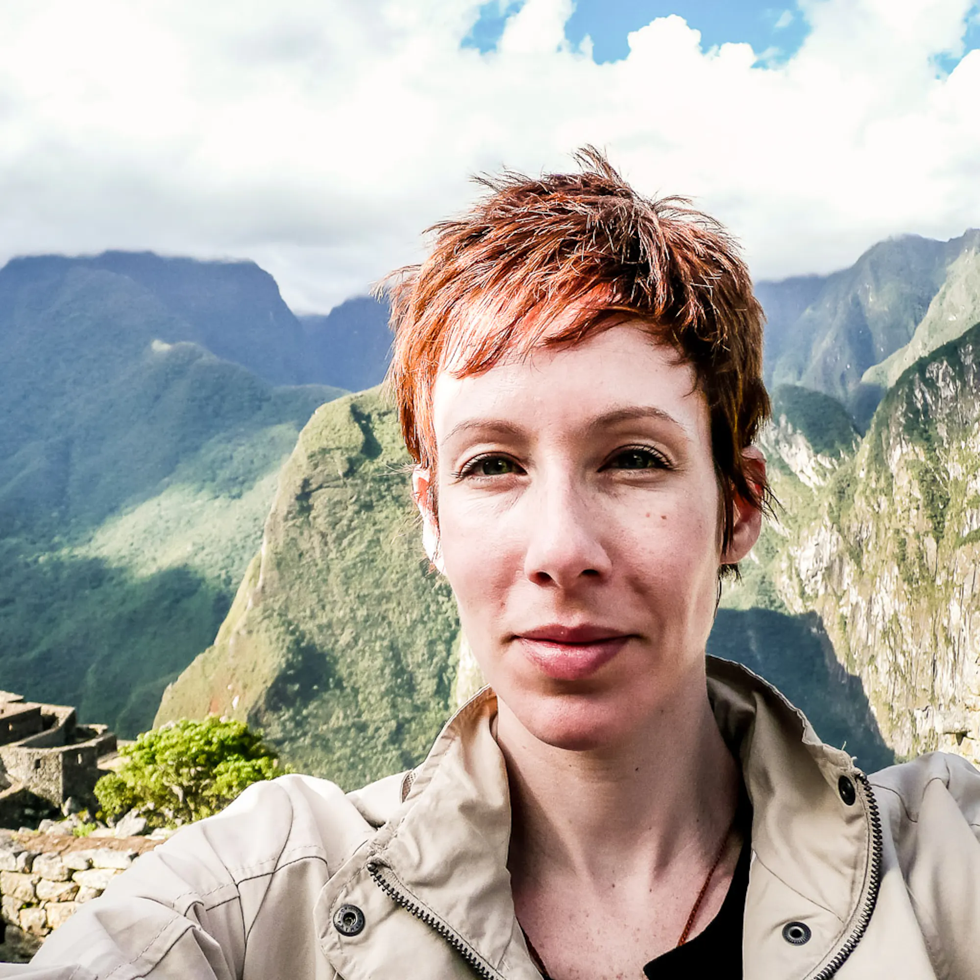 A close-up of a woman with short hair wearing a khaki jacket standing in front of moss-covered mountains.