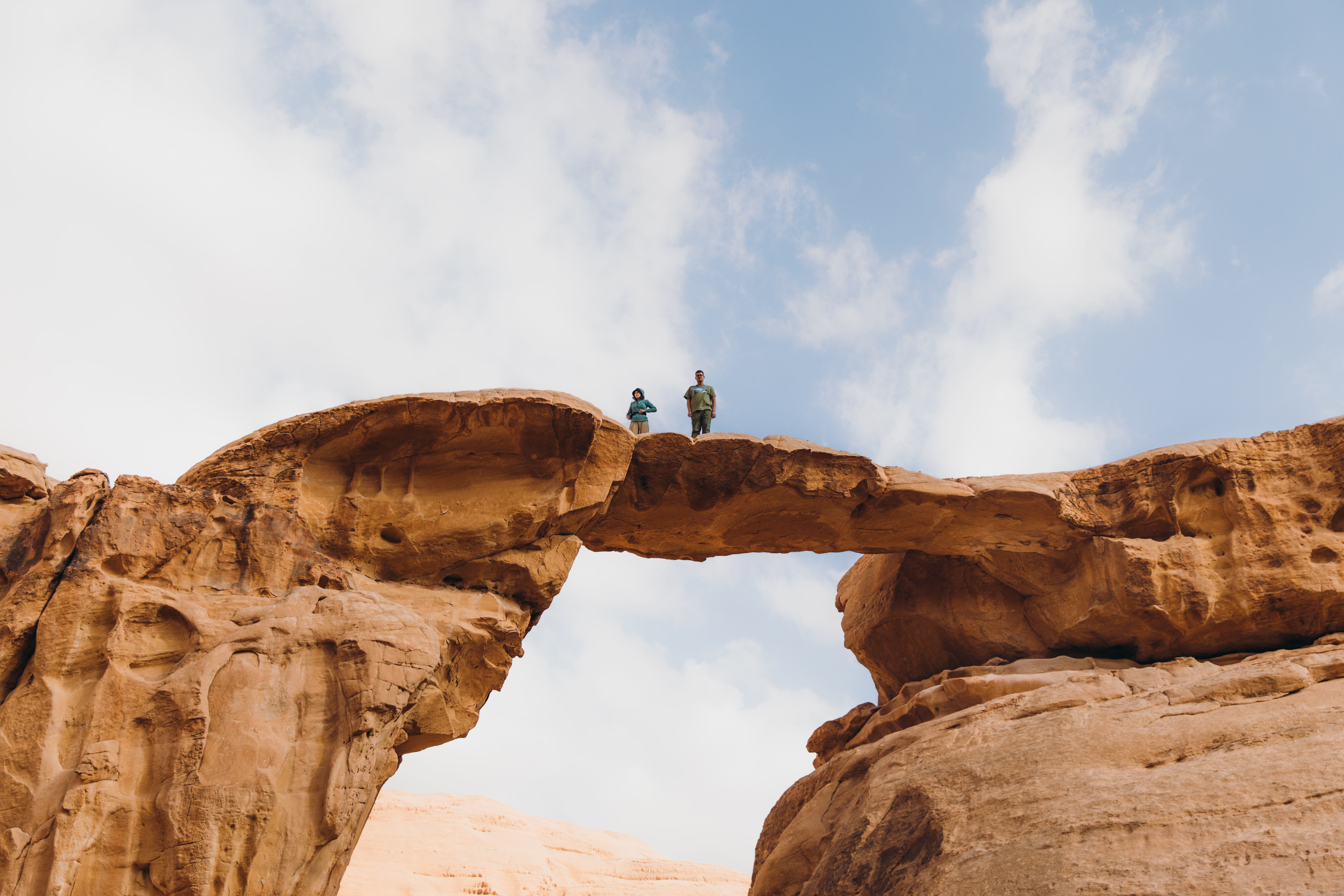 Two figures stand atop a sandstone arch overlooking the sweeping red desert of Wadi Rum.