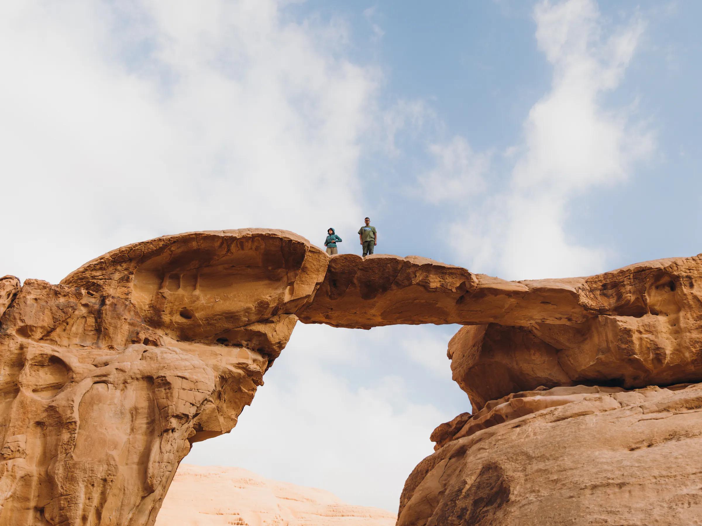 Two figures stand atop a sandstone arch overlooking the sweeping red desert of Wadi Rum.