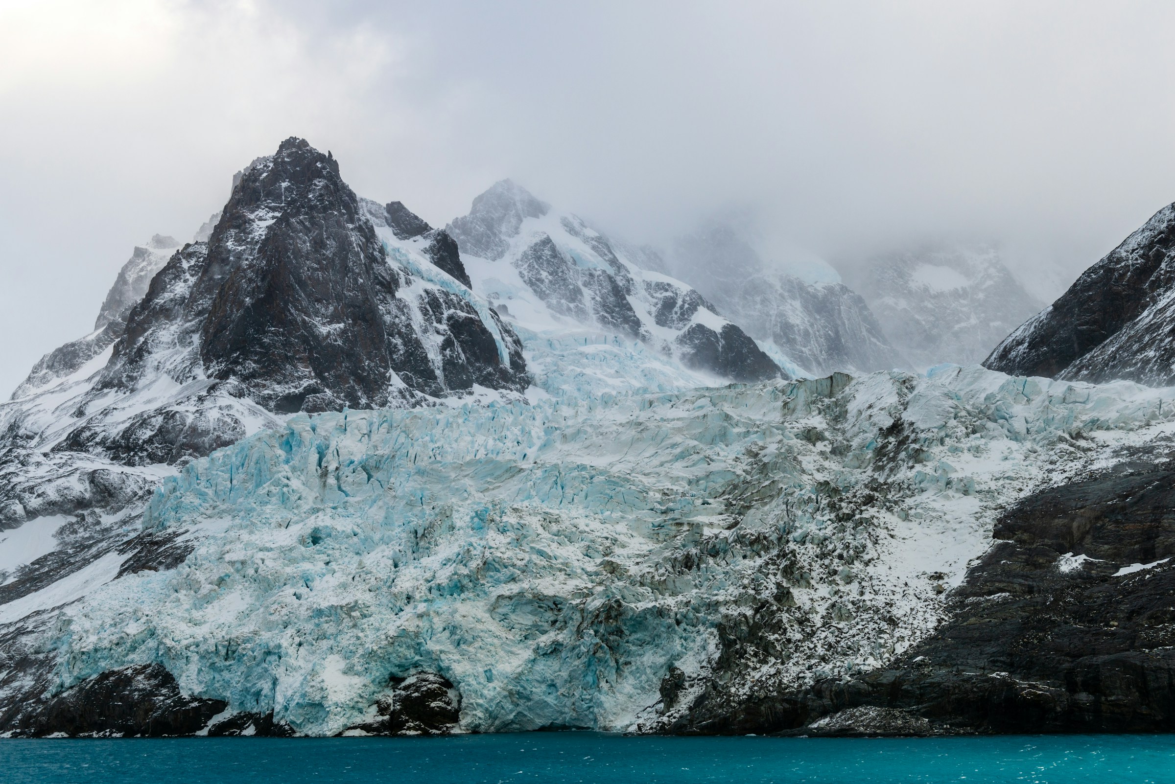 Jagged snow-covered peaks of South Georgia rise steeply above dark glacial waters.