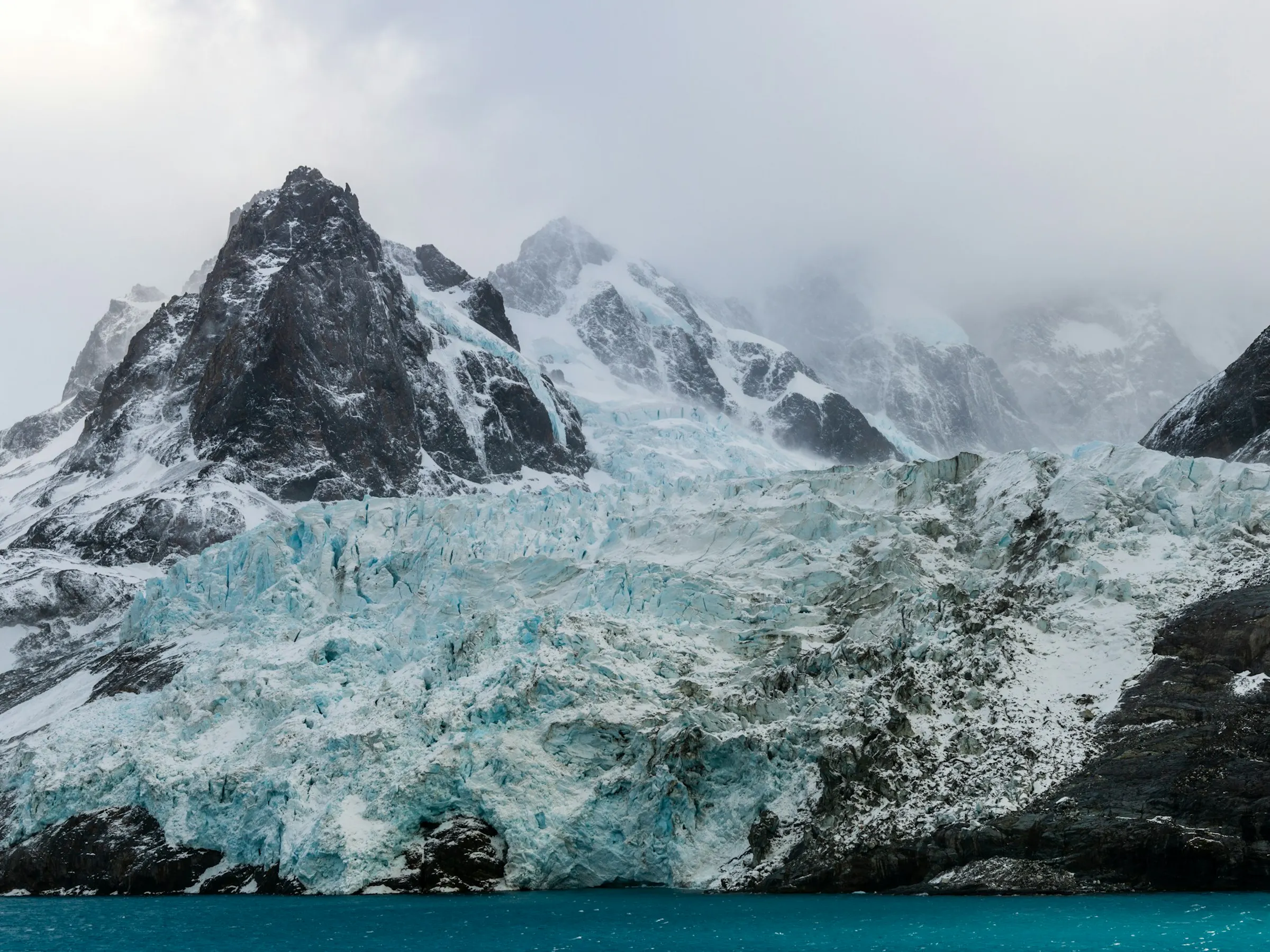 Jagged snow-covered peaks of South Georgia rise steeply above dark glacial waters.