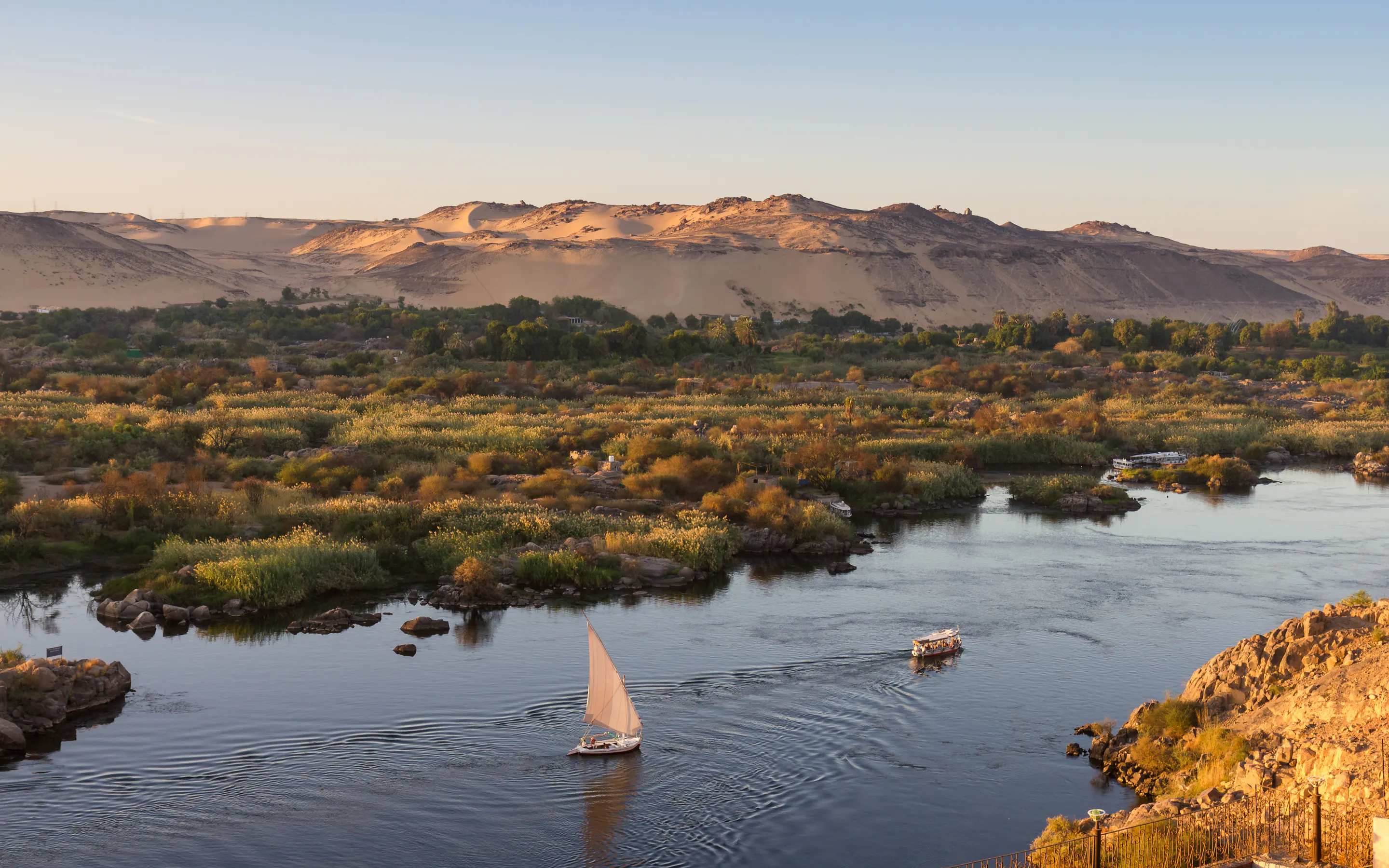 A felucca sailboat glides along the calm Nile River at Aswan with rocky banks in the background.