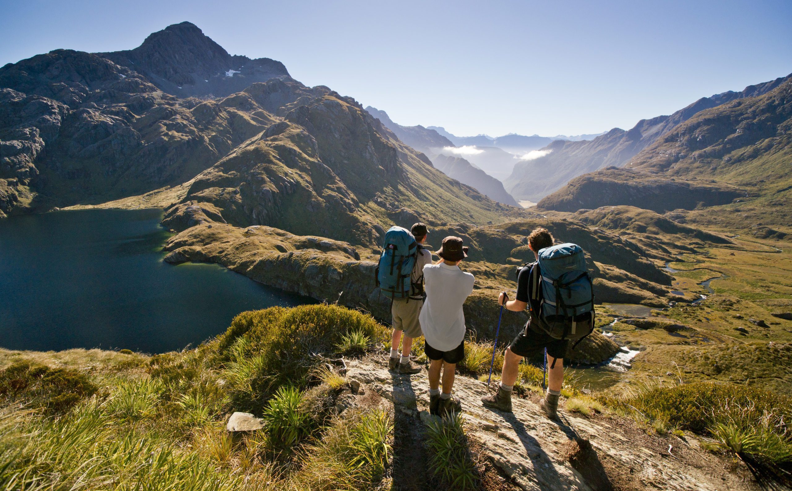 A hiker traverses a rugged alpine ridge on the Routeburn Track with misty Fiordland peaks beyond.