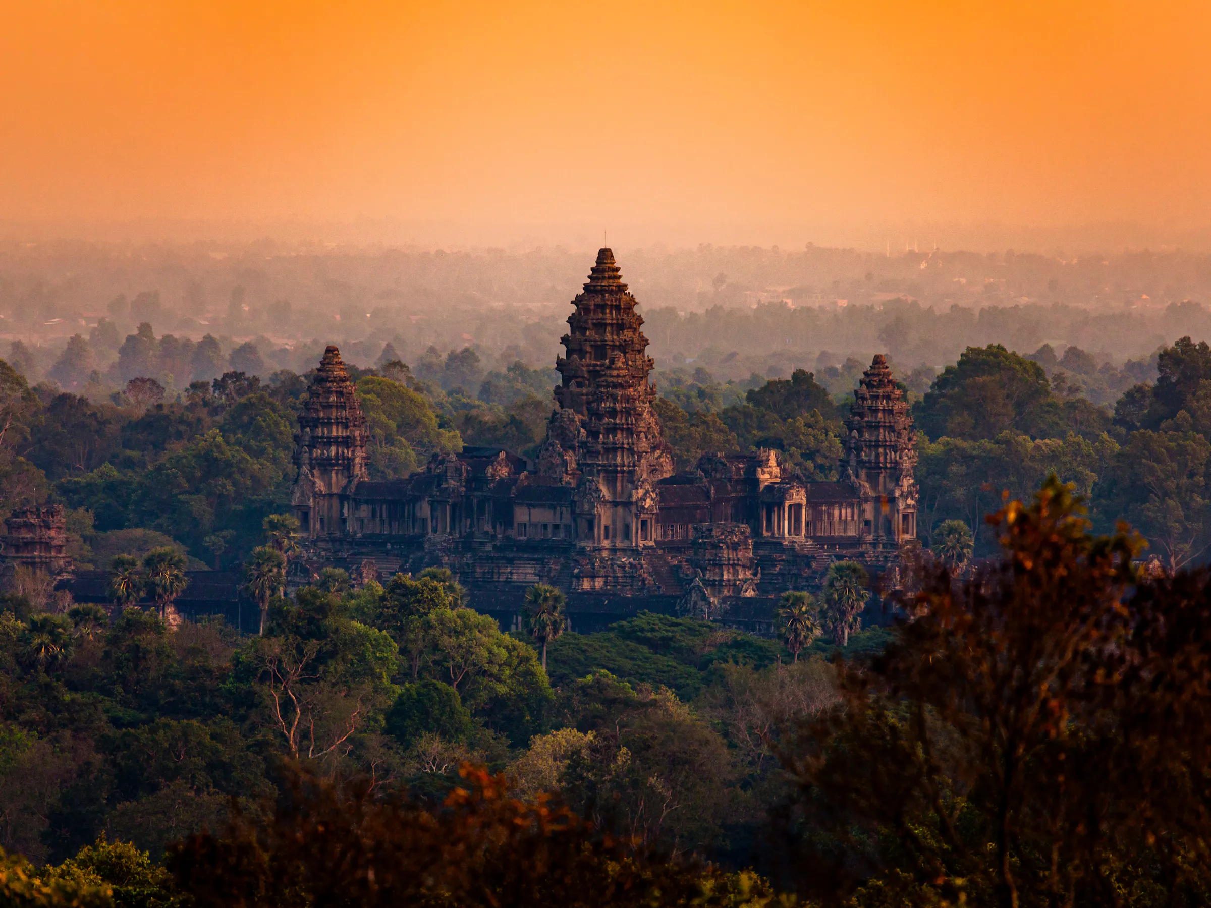 The temple towers of Angkor Wat are reflected in a still pool at dusk under a deep orange sky.