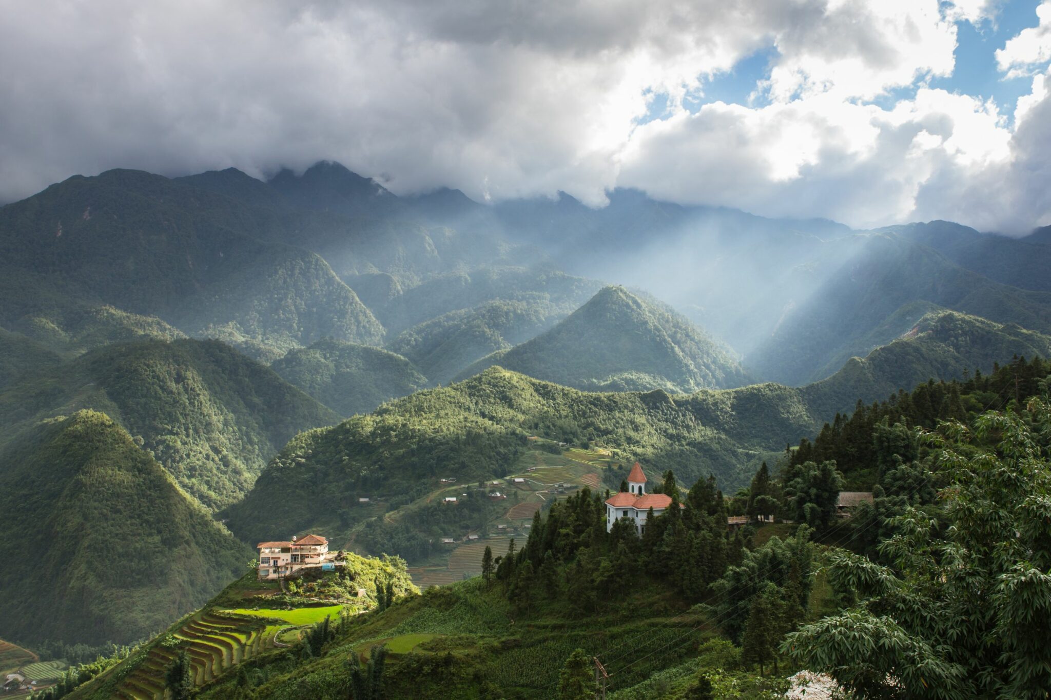 Mist-shrouded mountains rise behind a forested valley and distant terraced hillsides in Sapa.