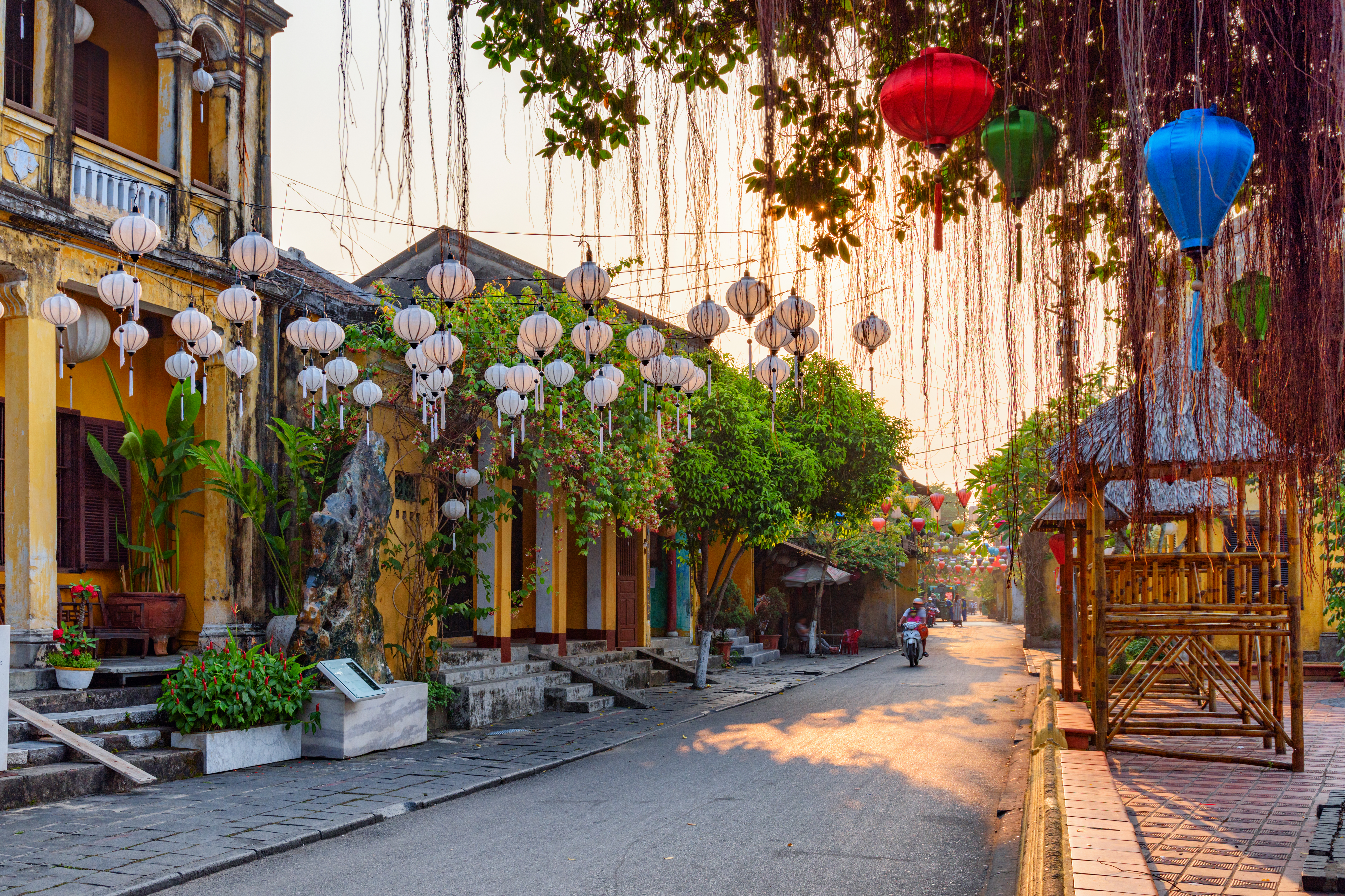 Silk lanterns in vivid red, yellow, and blue hang above a golden-walled street in Hoi An at dusk.