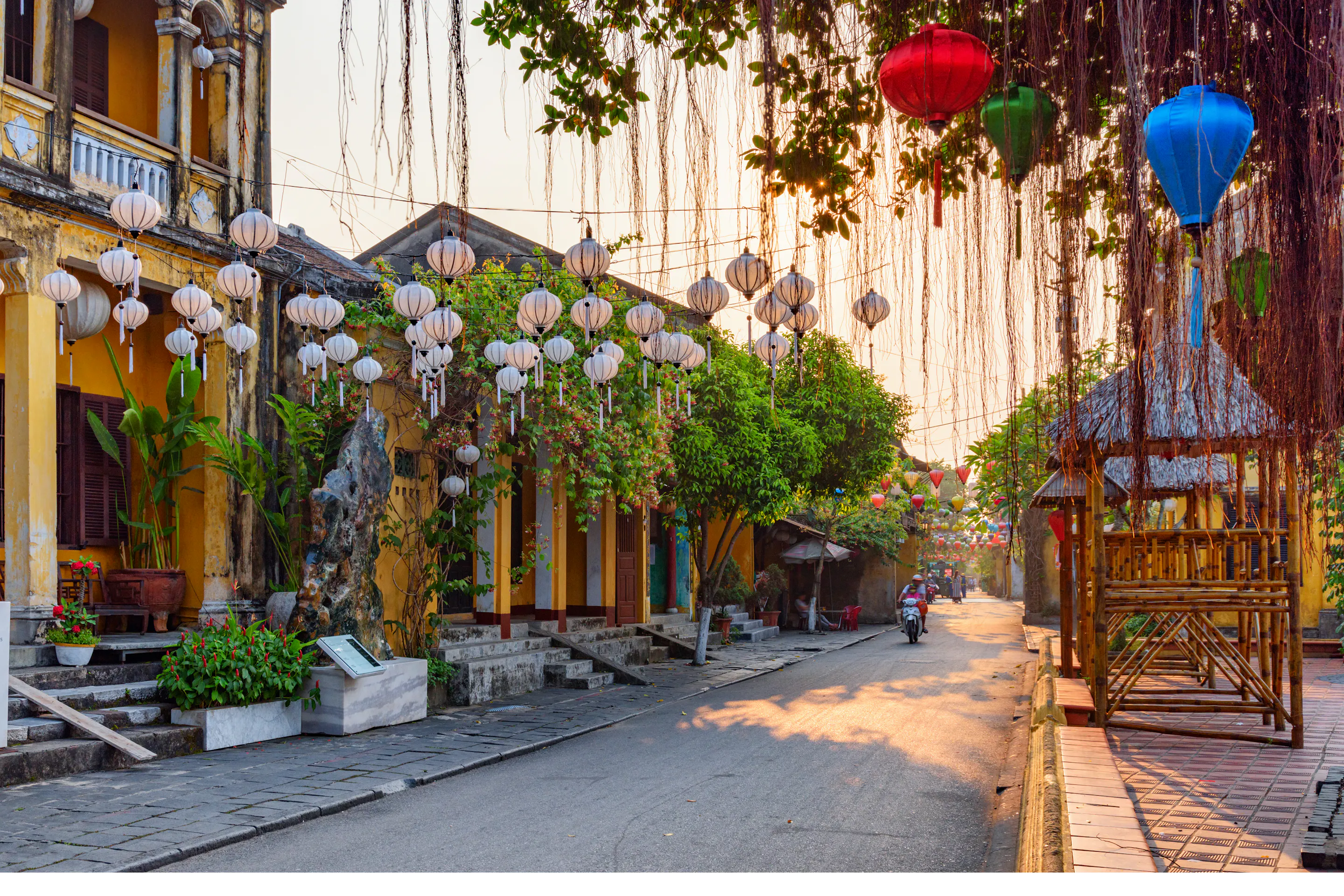 Silk lanterns in vivid red, yellow, and blue hang above a golden-walled street in Hoi An at dusk.