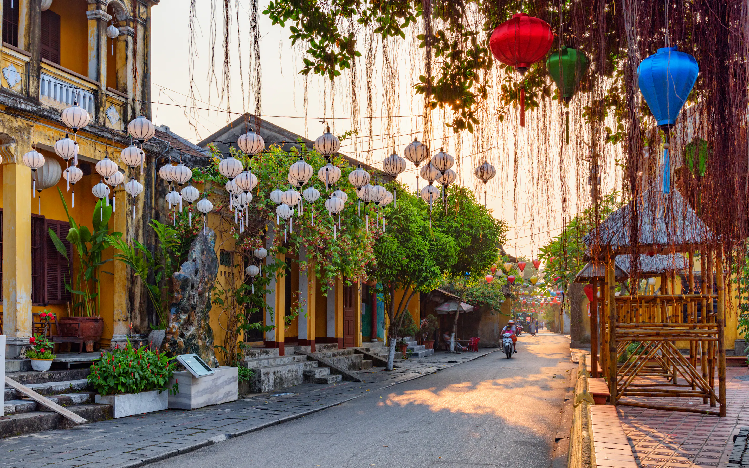 Silk lanterns in vivid red, yellow, and blue hang above a golden-walled street in Hoi An at dusk.