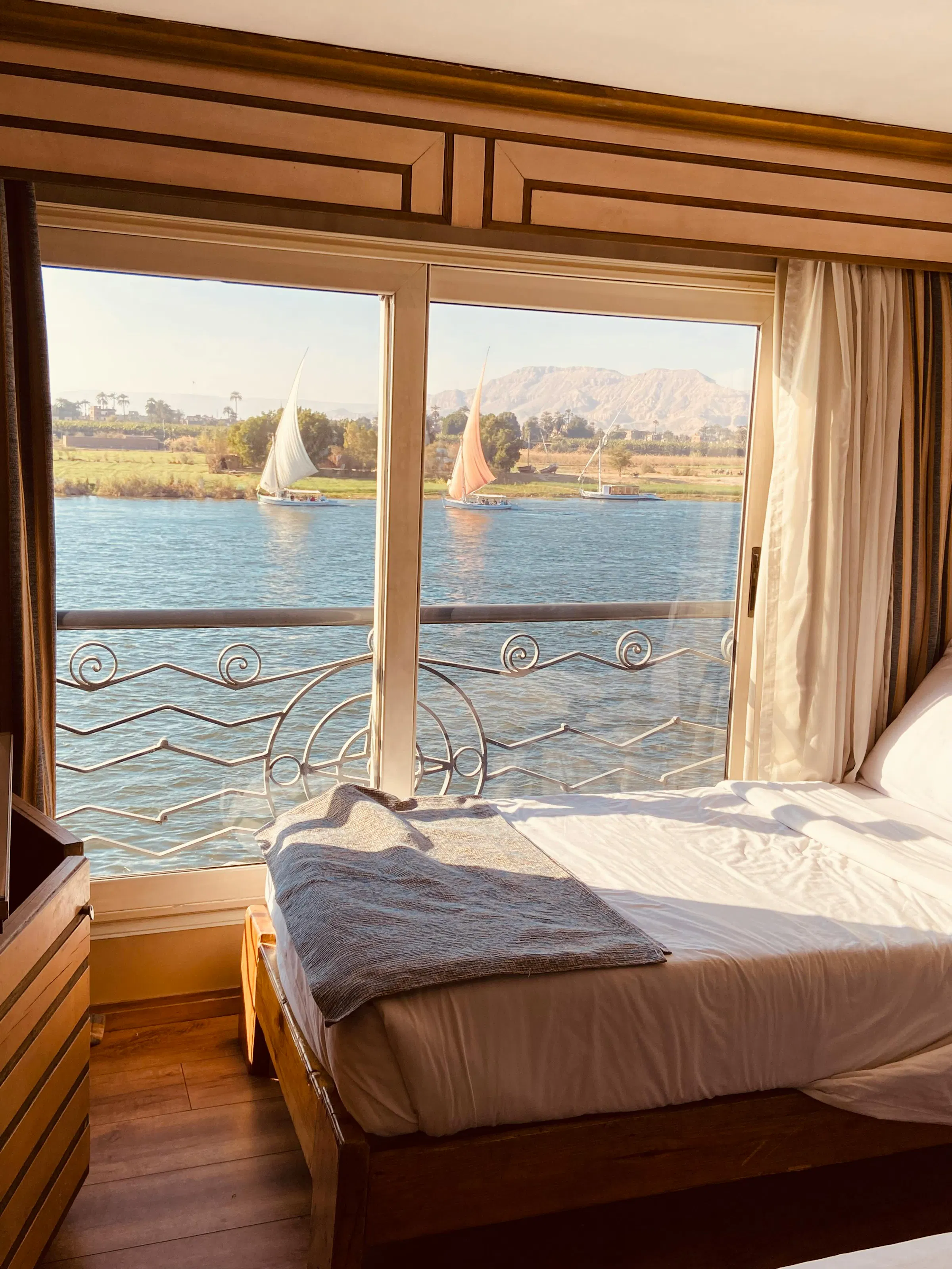 A bedroom with wide windows frames a serene view of sailboats on the Nile River at Aswan.