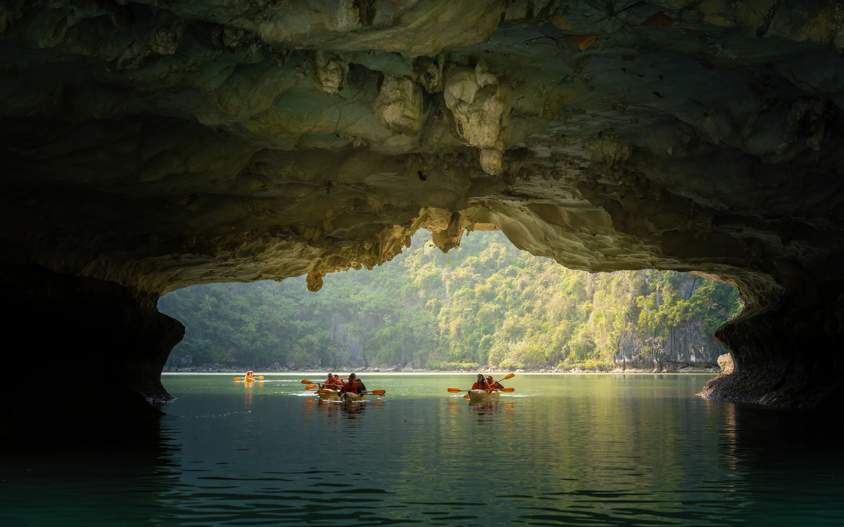 A kayaker paddles through calm jade-green waters between towering limestone karsts in Ha Long Bay.