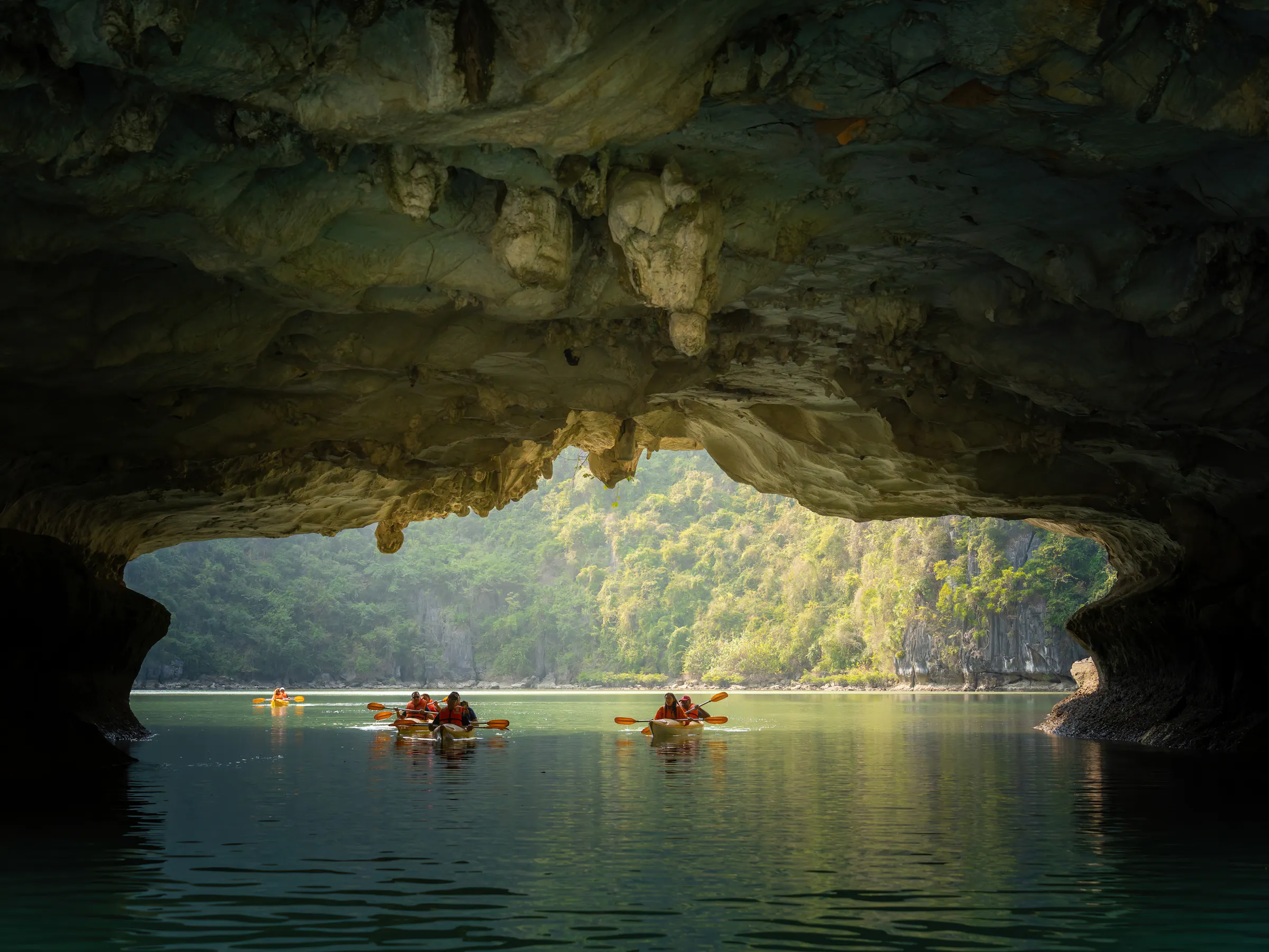 A kayaker paddles through calm jade-green waters between towering limestone karsts in Ha Long Bay.