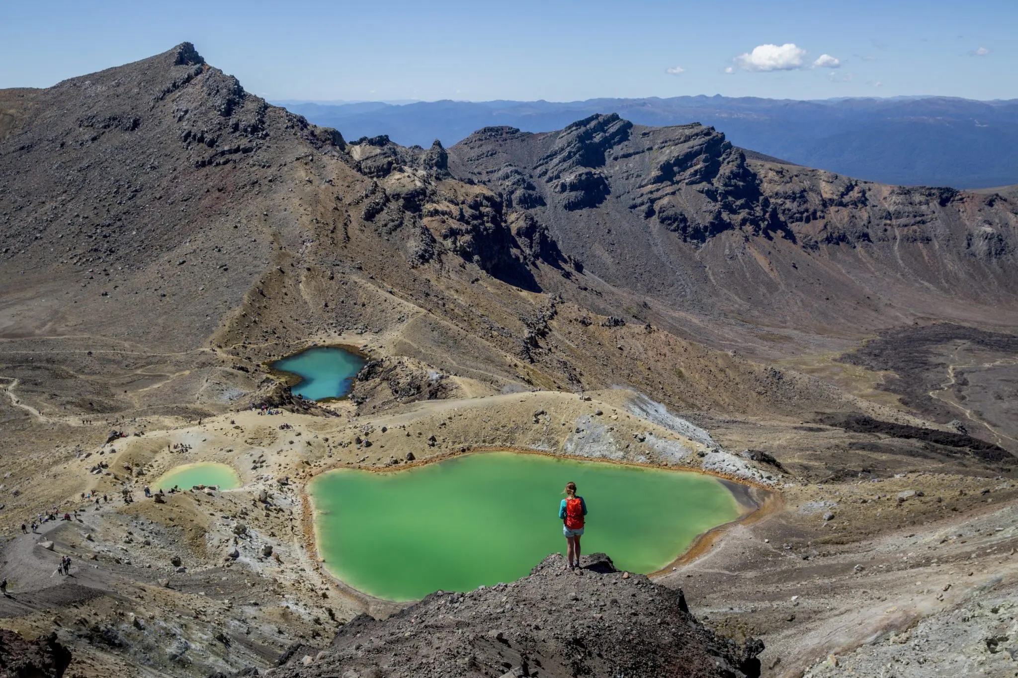 Two hikers stand beside an emerald crater lake on the Tongariro Alpine Crossing with peaks beyond.
