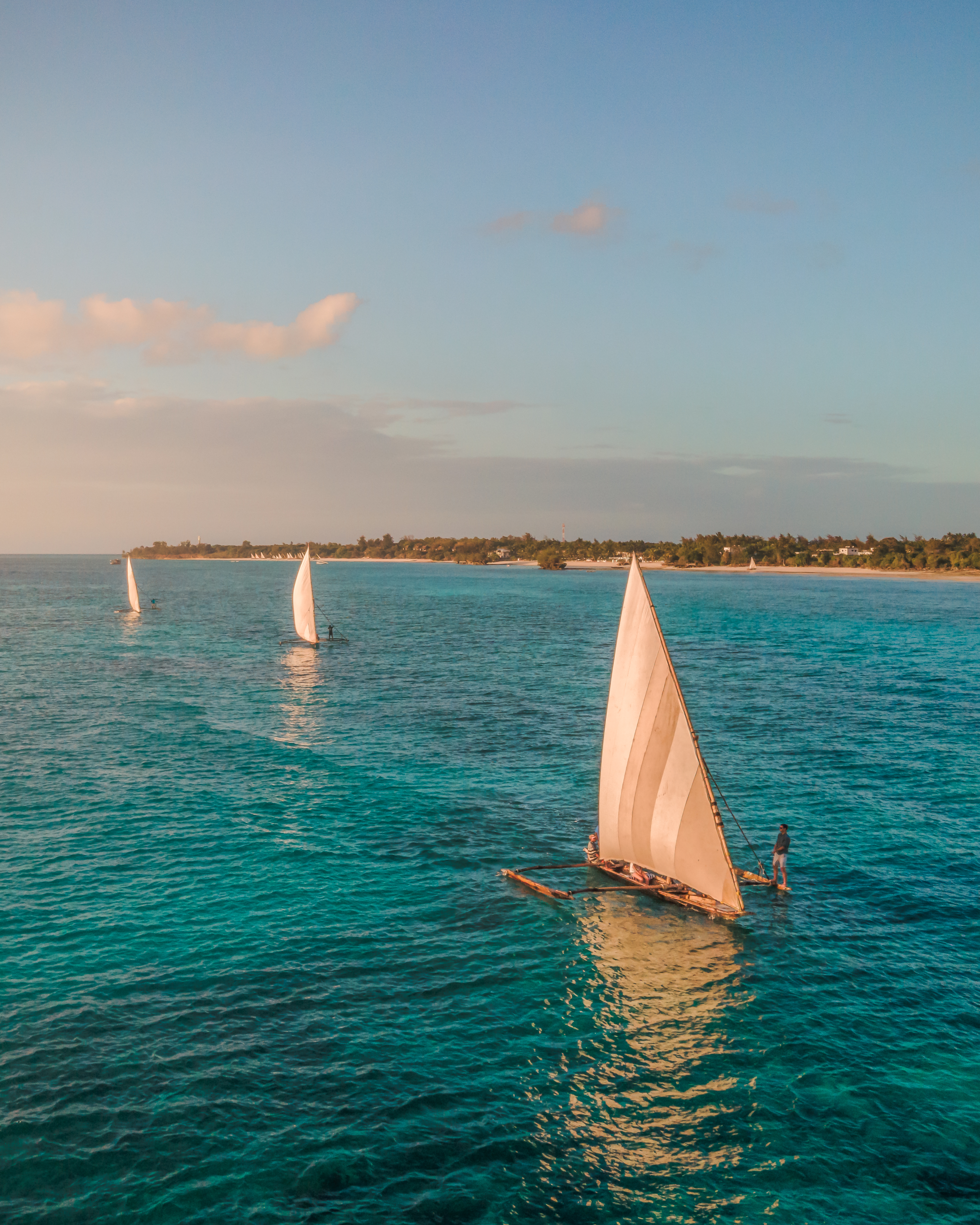 A traditional dhow sailboat glides across calm teal water with a vivid orange sunset on the horizon.