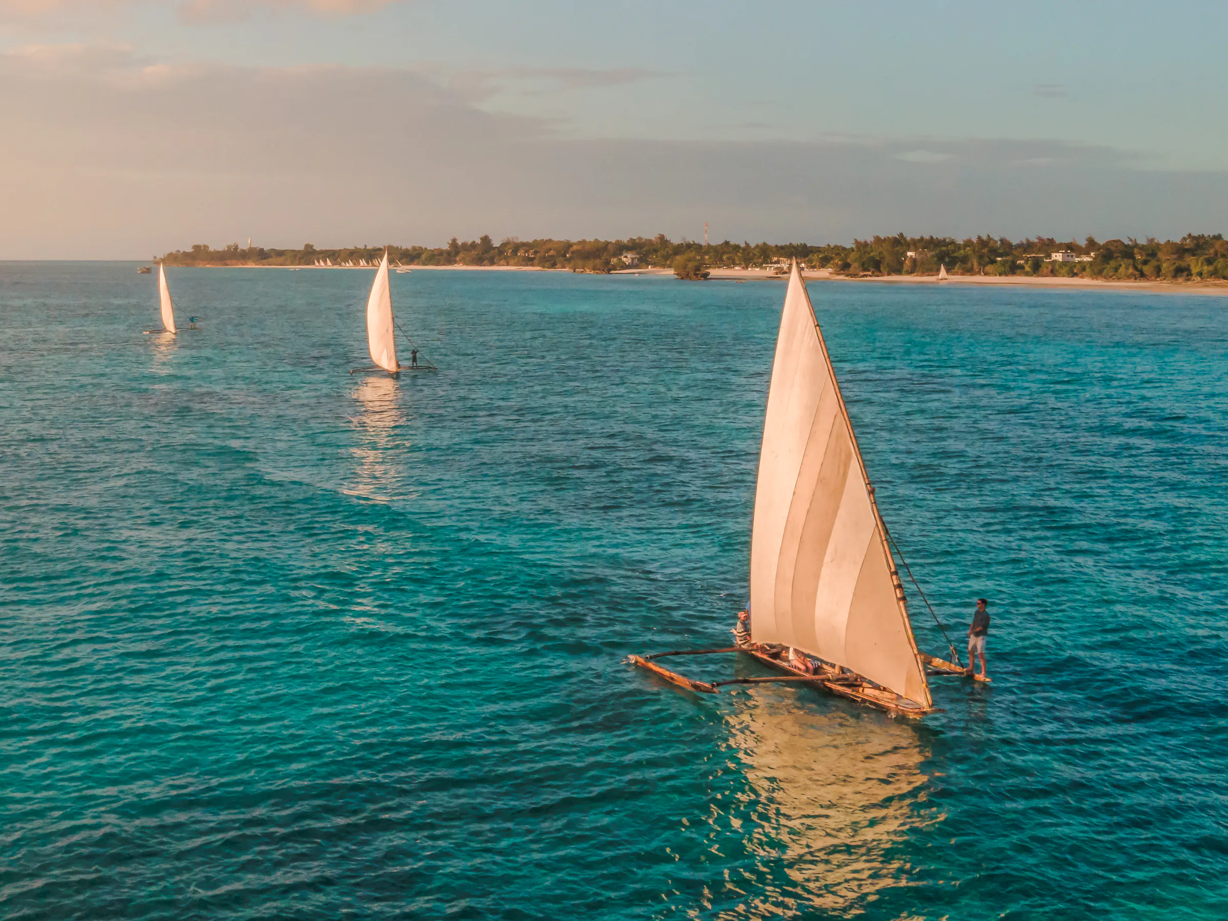 A traditional dhow sailboat glides across calm teal water with a vivid orange sunset on the horizon.