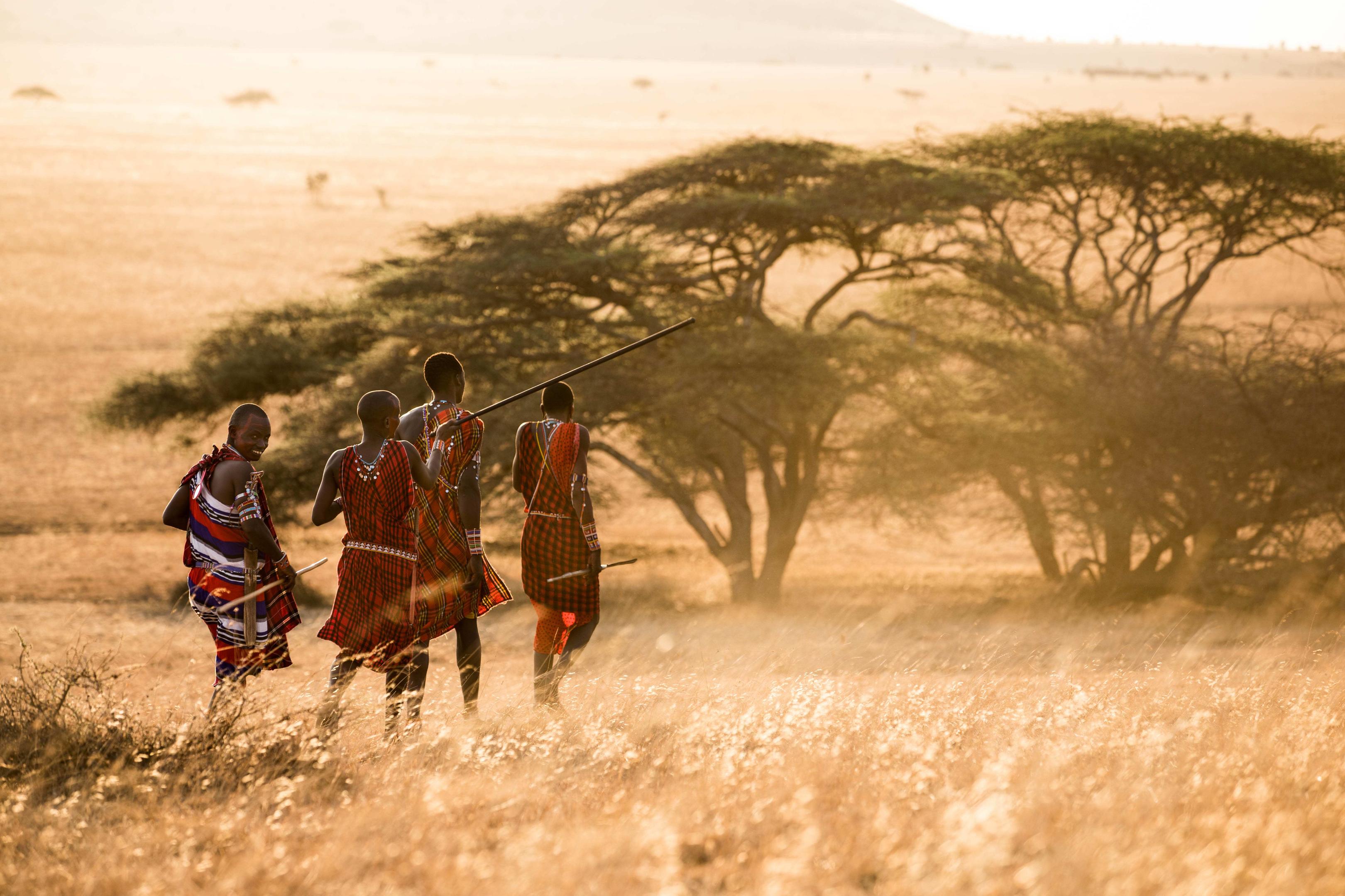 Three Maasai figures in red robes walk through a dusty golden landscape beneath wide acacia trees.