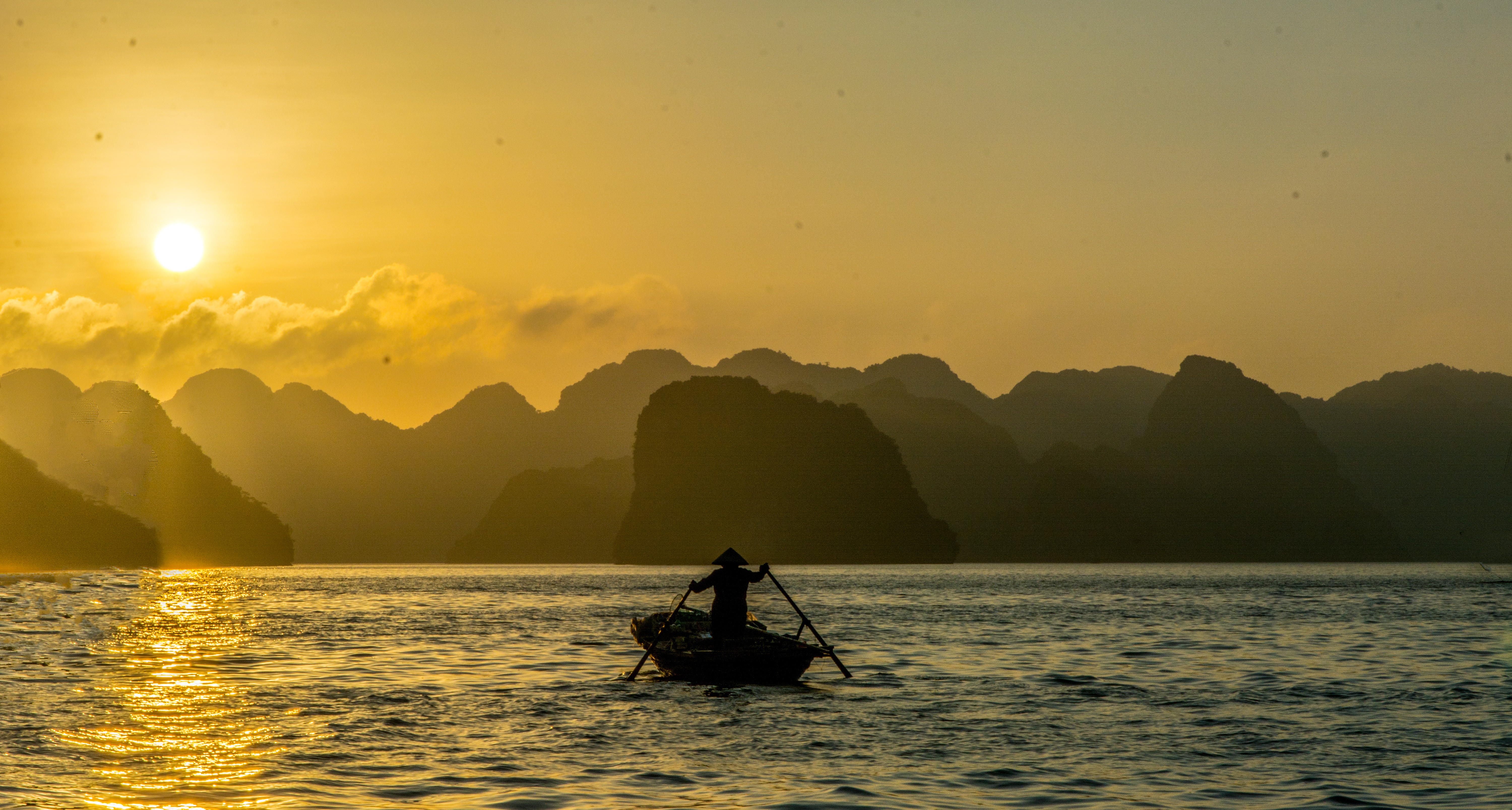 A traditional junk boat sails between towering limestone karsts at sunset in Ha Long Bay.