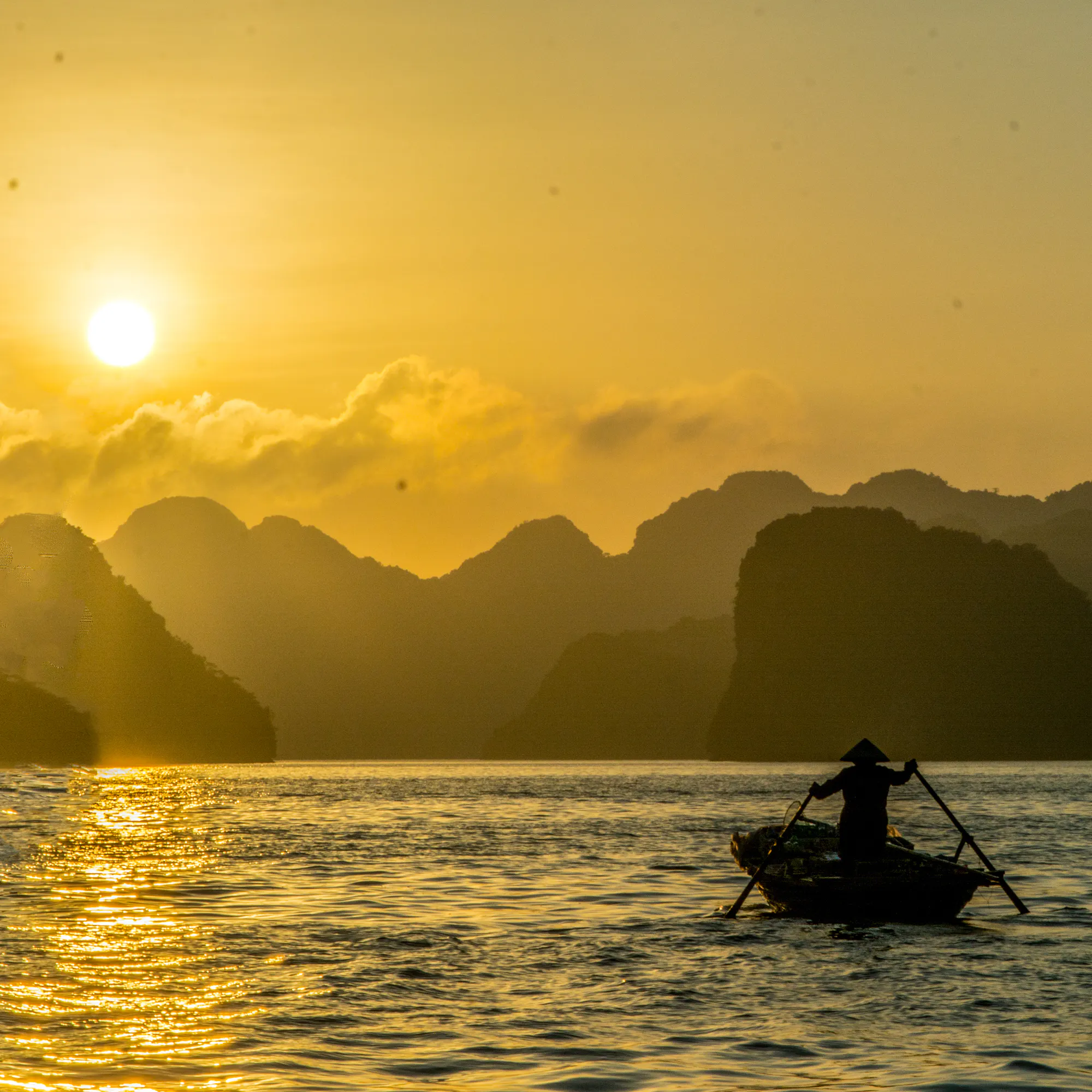 A traditional junk boat sails between towering limestone karsts at sunset in Ha Long Bay.