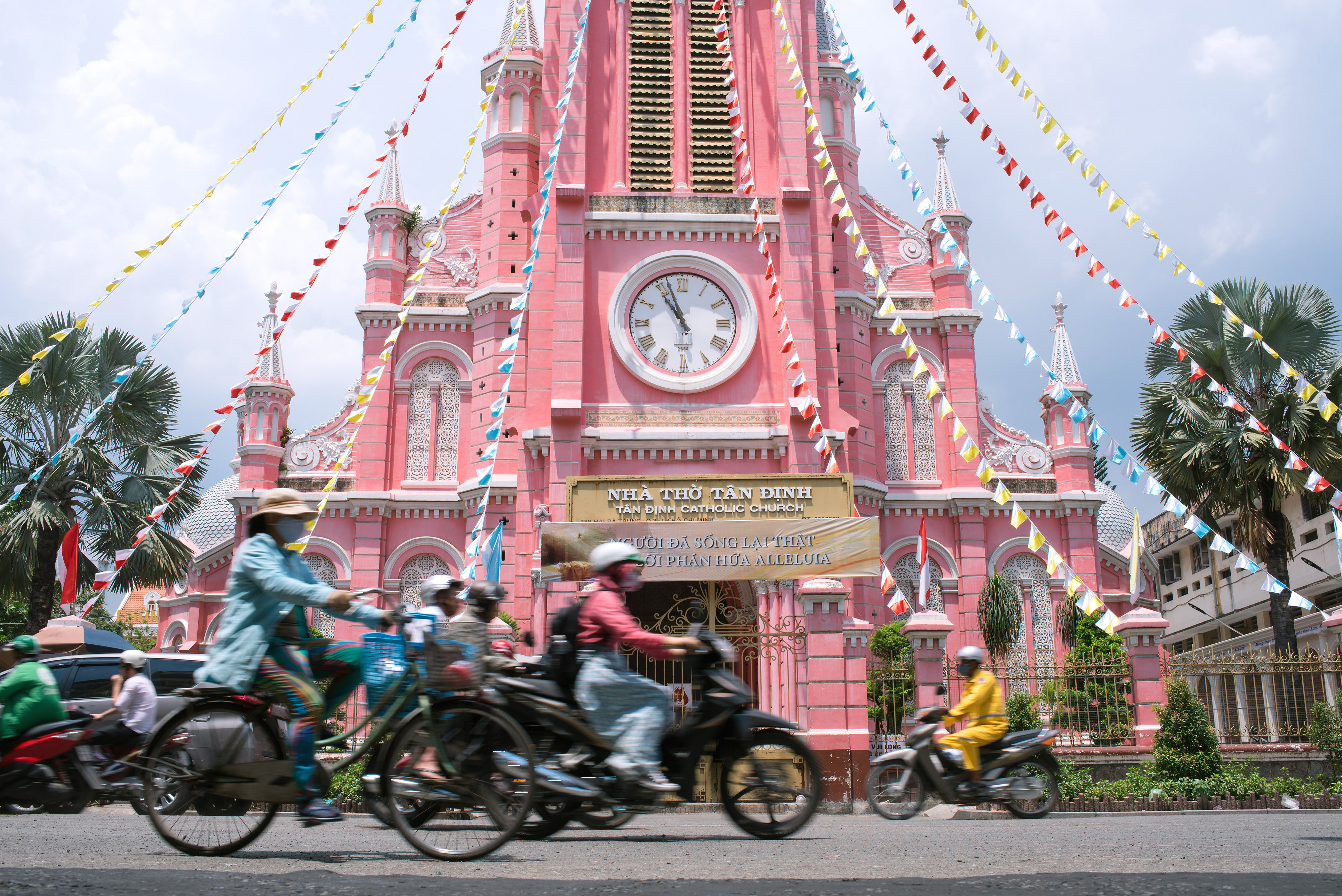 A busy Ho Chi Minh City street is filled with motorcycles beside a pink colonial clock tower.