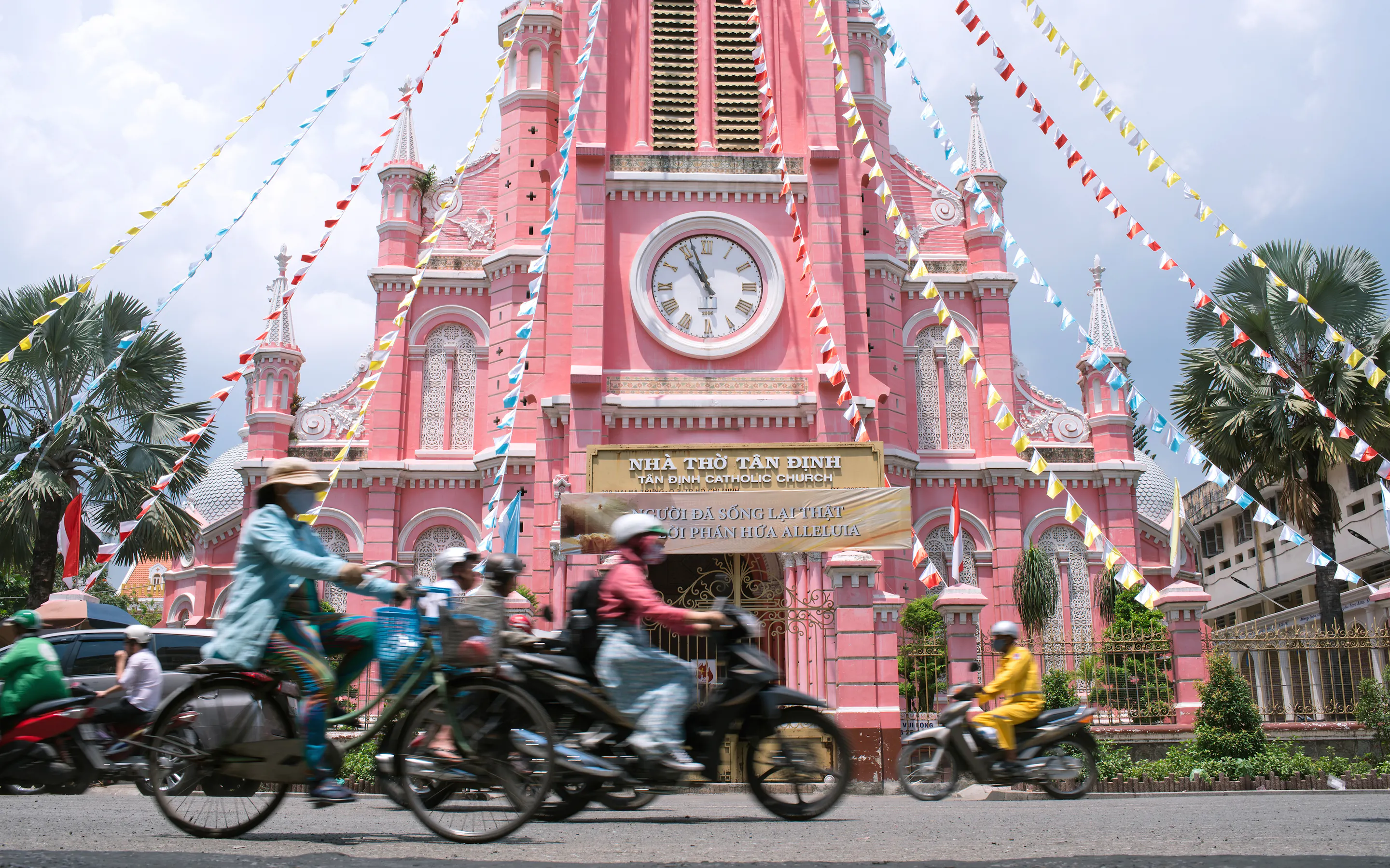 A busy Ho Chi Minh City street is filled with motorcycles beside a pink colonial clock tower.