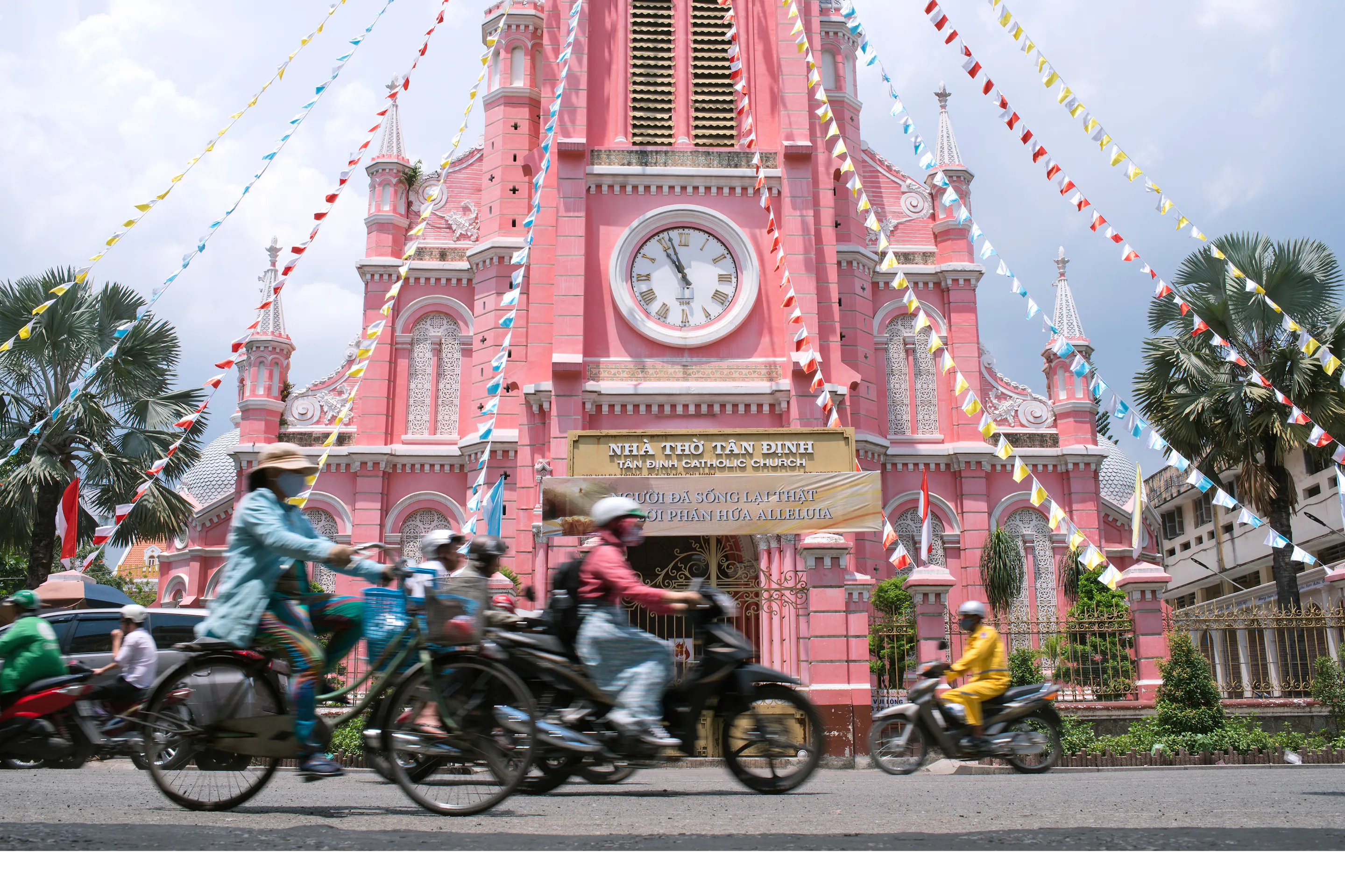 A busy Ho Chi Minh City street is filled with motorcycles beside a pink colonial clock tower.