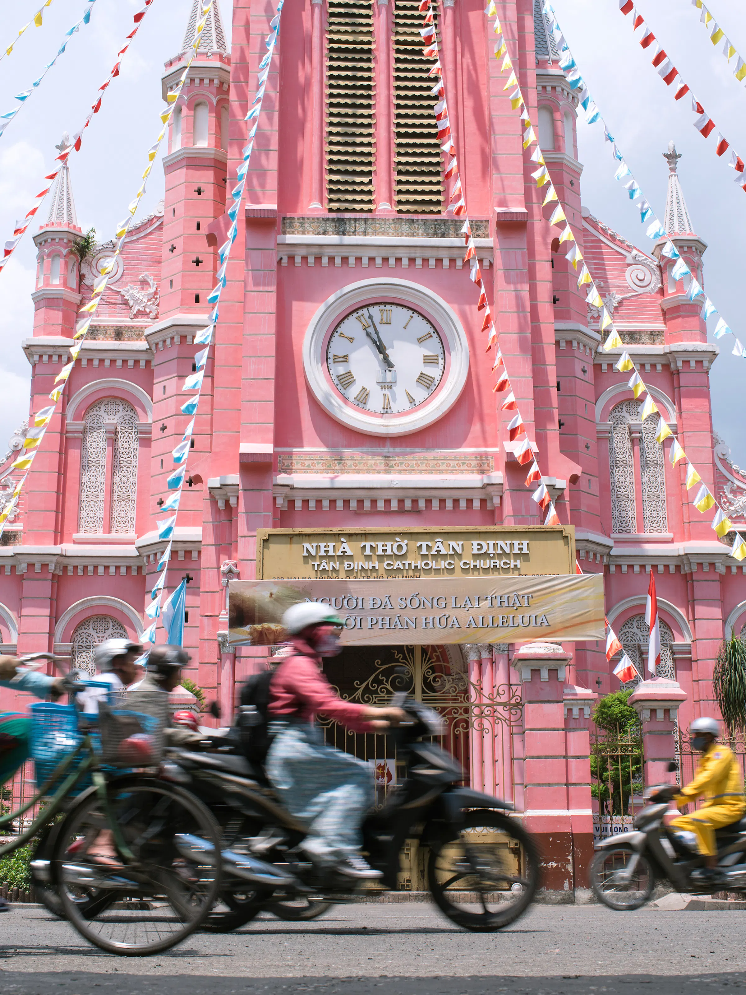 A busy Ho Chi Minh City street is filled with motorcycles beside a pink colonial clock tower.