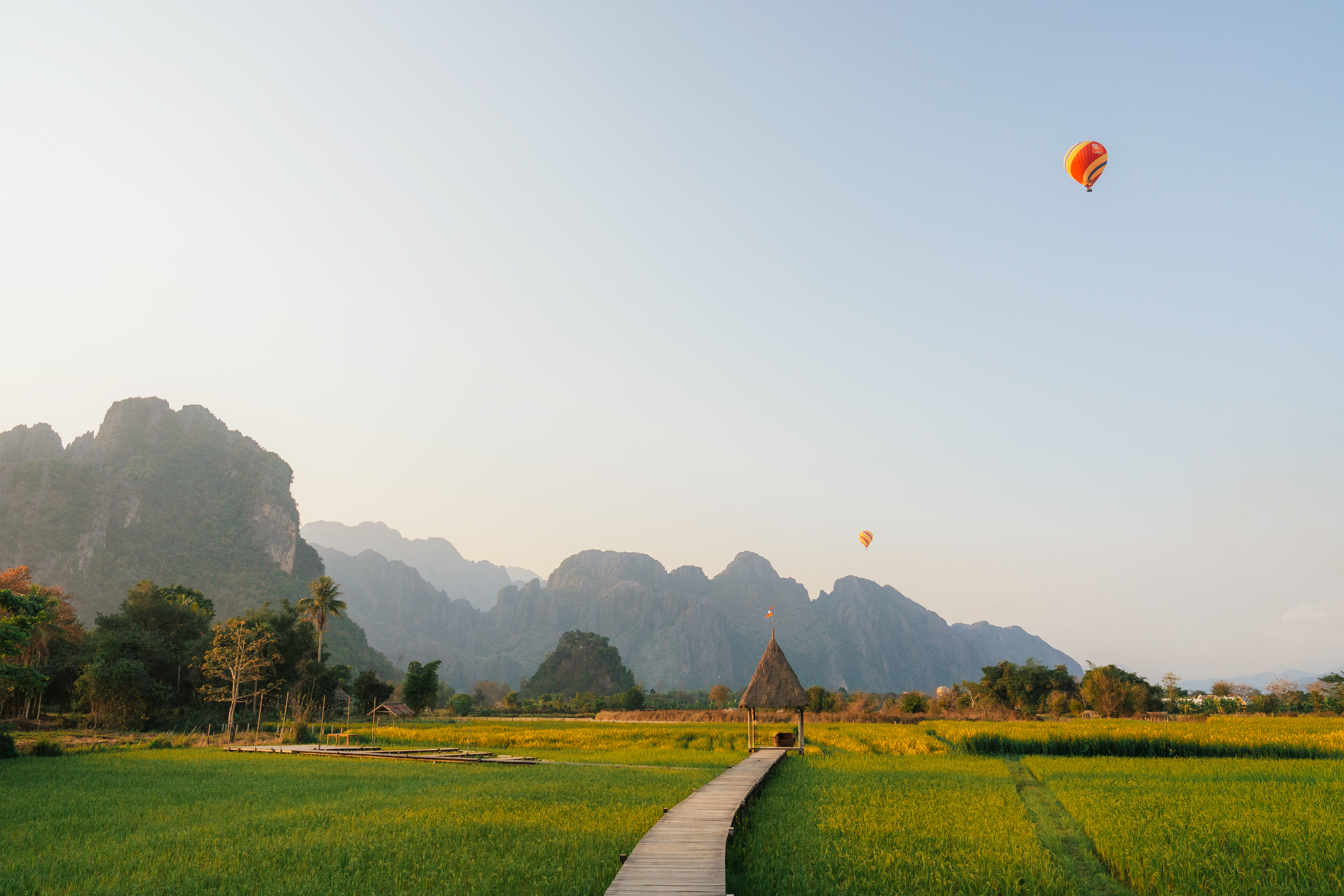 A red hot-air balloon floats above flat green fields with dramatic karst peaks in the background.