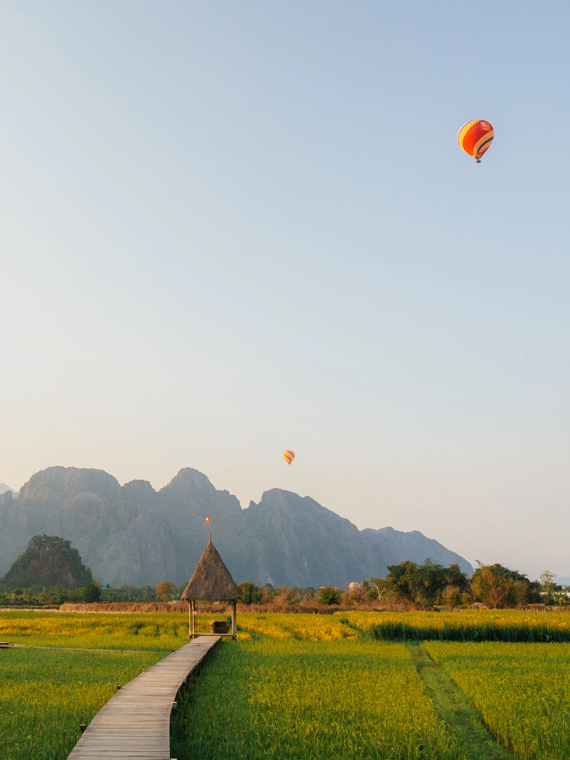 A red hot-air balloon floats above flat green fields with dramatic karst peaks in the background.