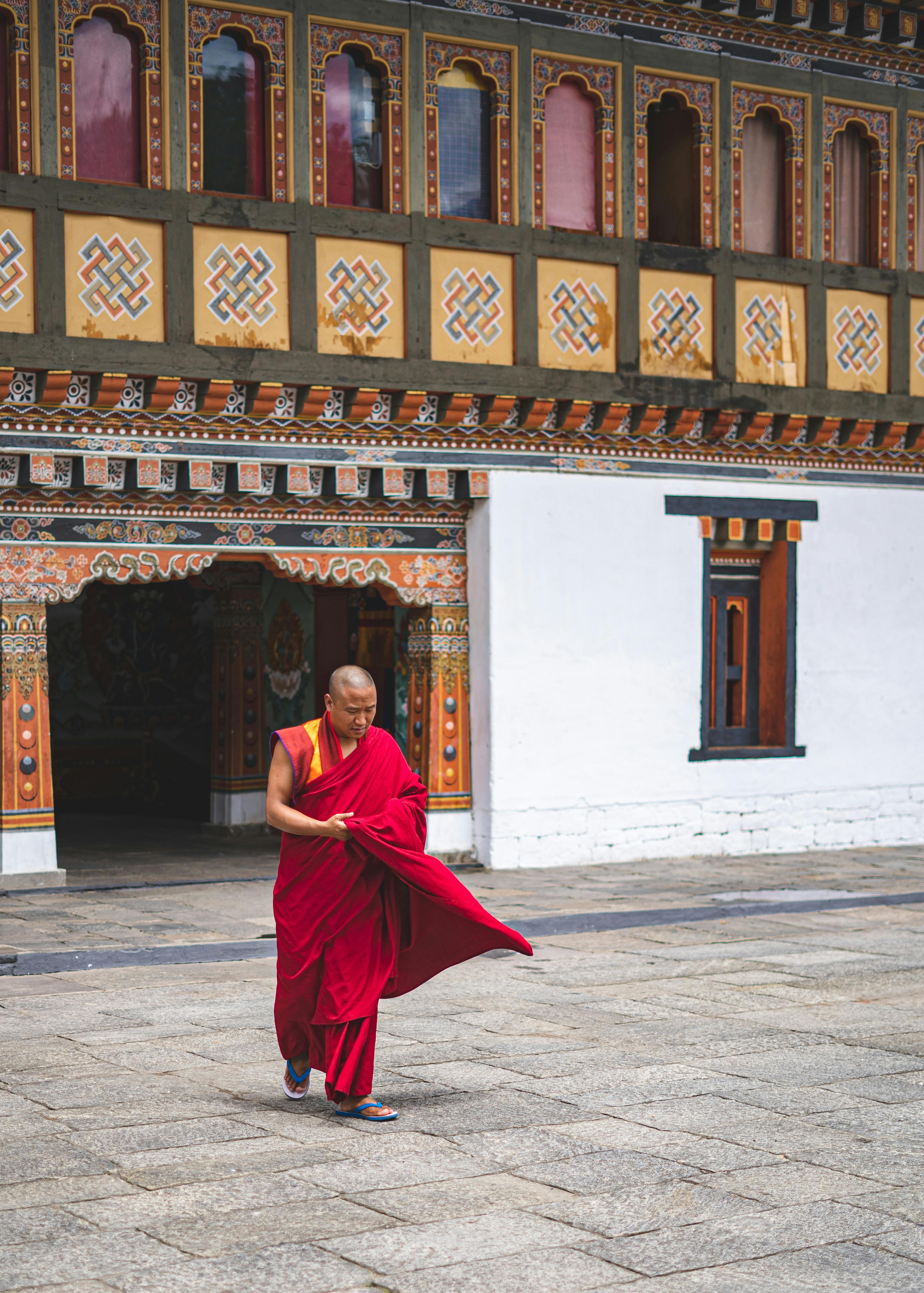 A monk in a red robe walks through the ornate white gateway of a traditional Bhutanese dzong.