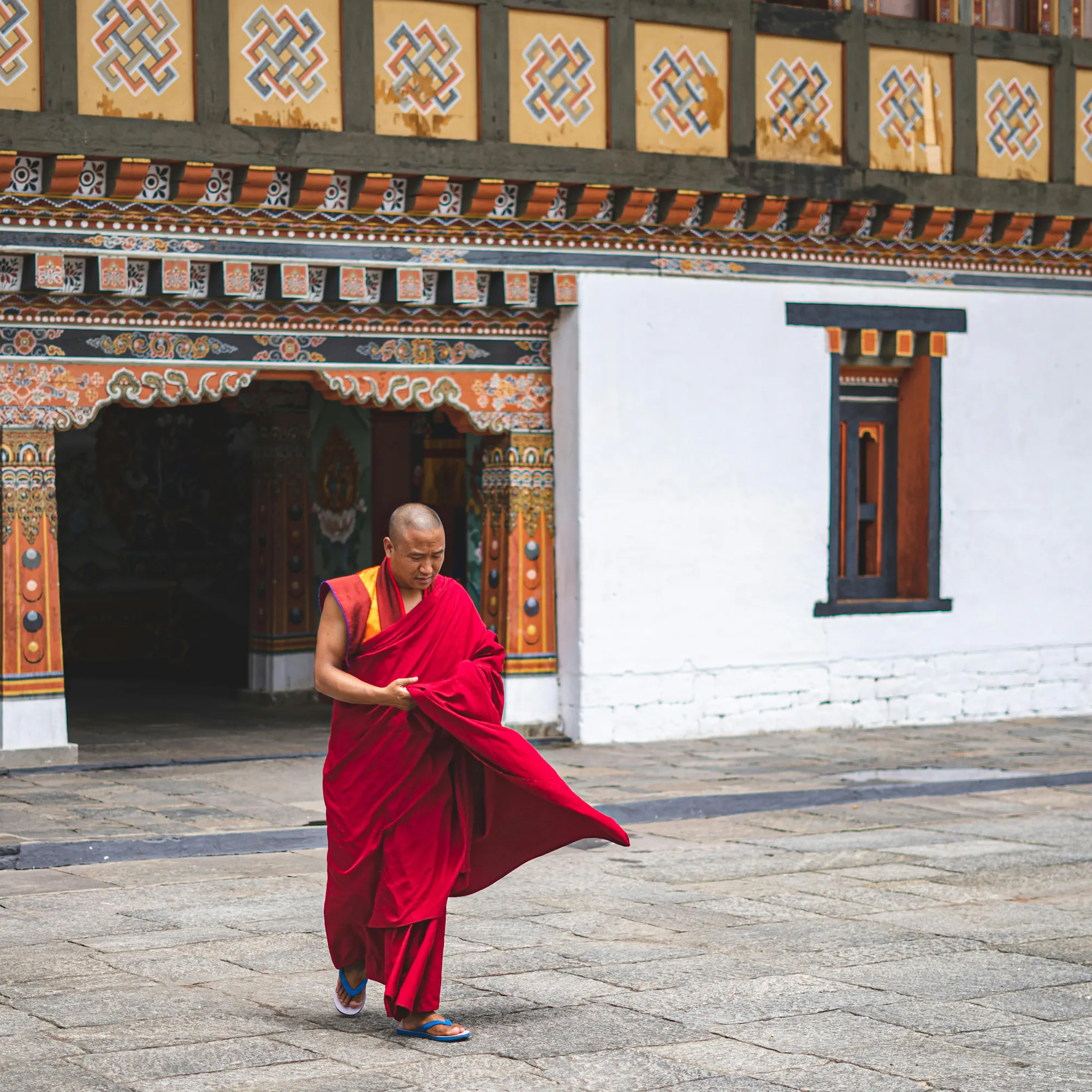 A monk in a red robe walks through the ornate white gateway of a traditional Bhutanese dzong.