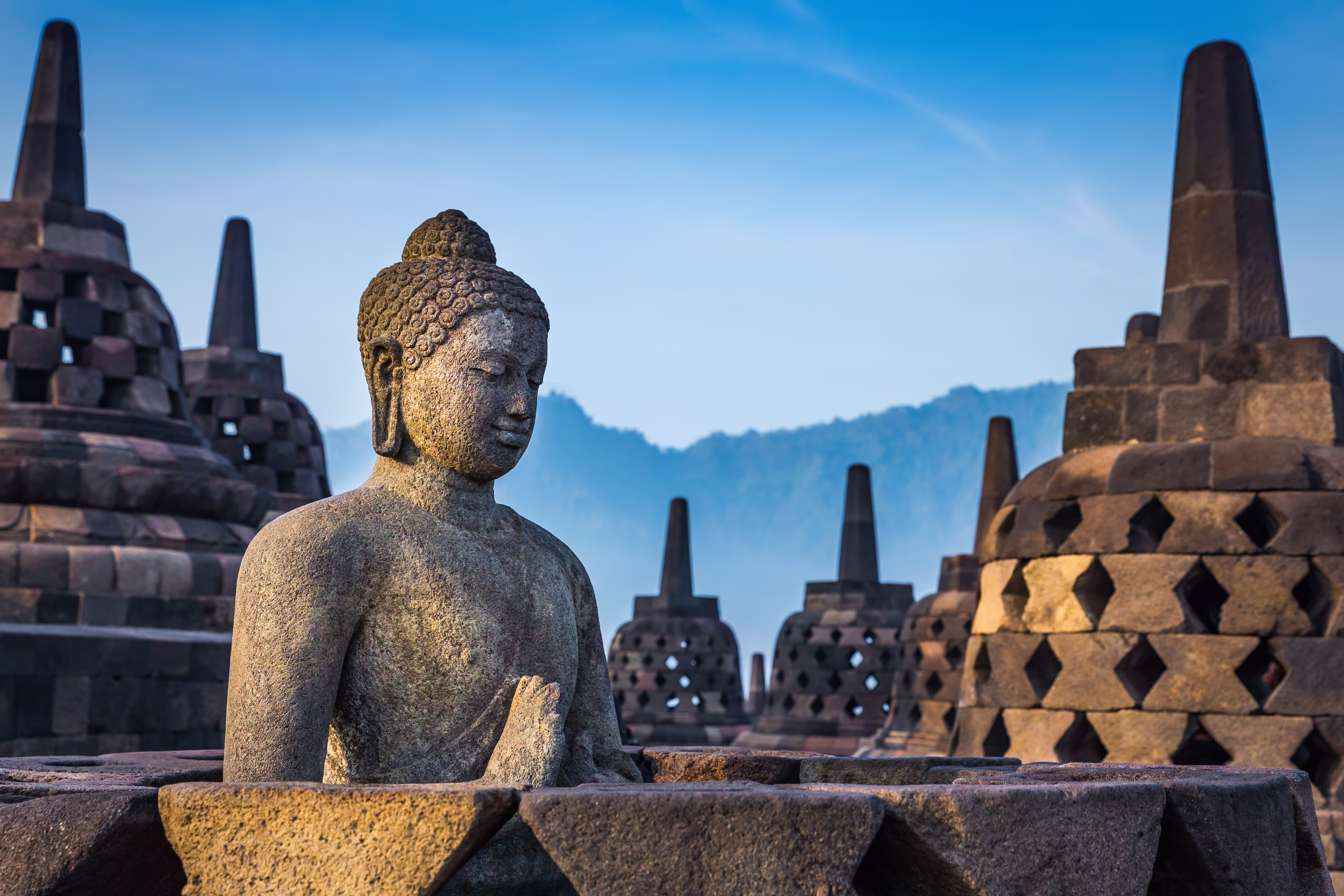 A large seated Buddha statue with intricate carvings sits before stupas and blue sky at Borobudur.
