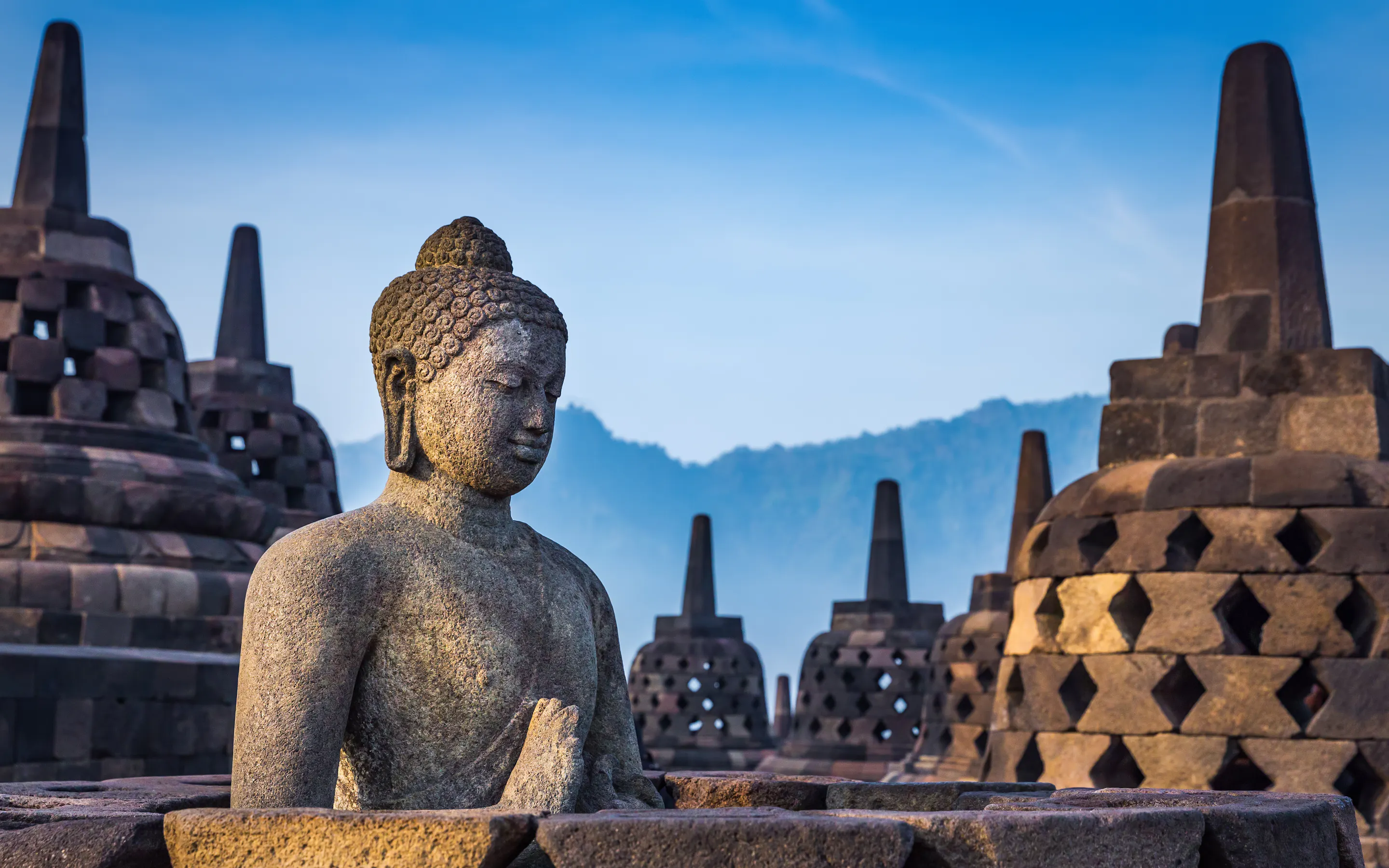A large seated Buddha statue with intricate carvings sits before stupas and blue sky at Borobudur.