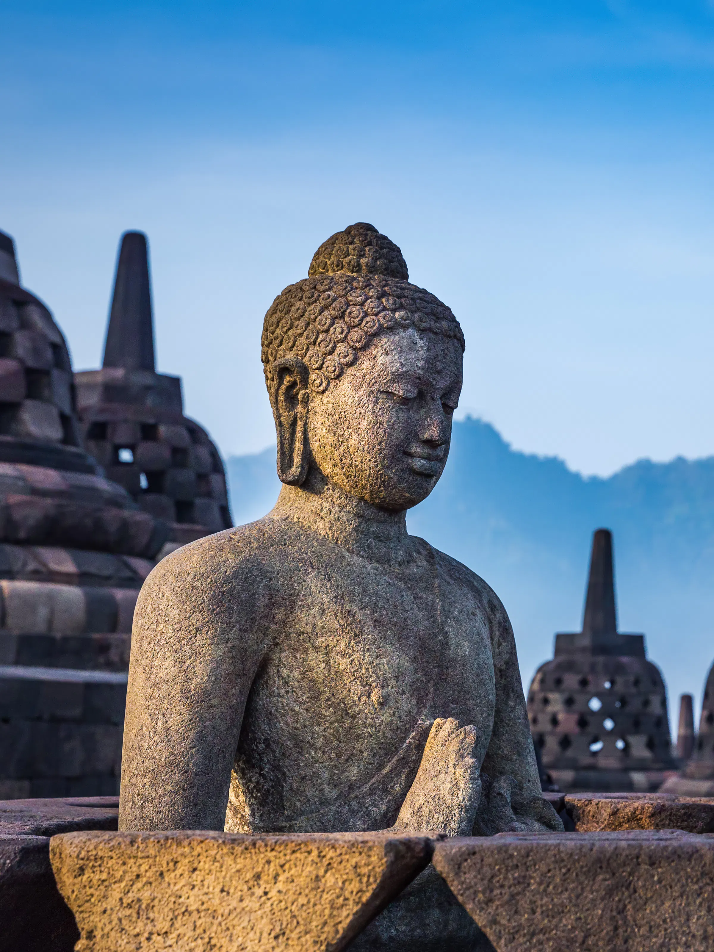 A large seated Buddha statue with intricate carvings sits before stupas and blue sky at Borobudur.
