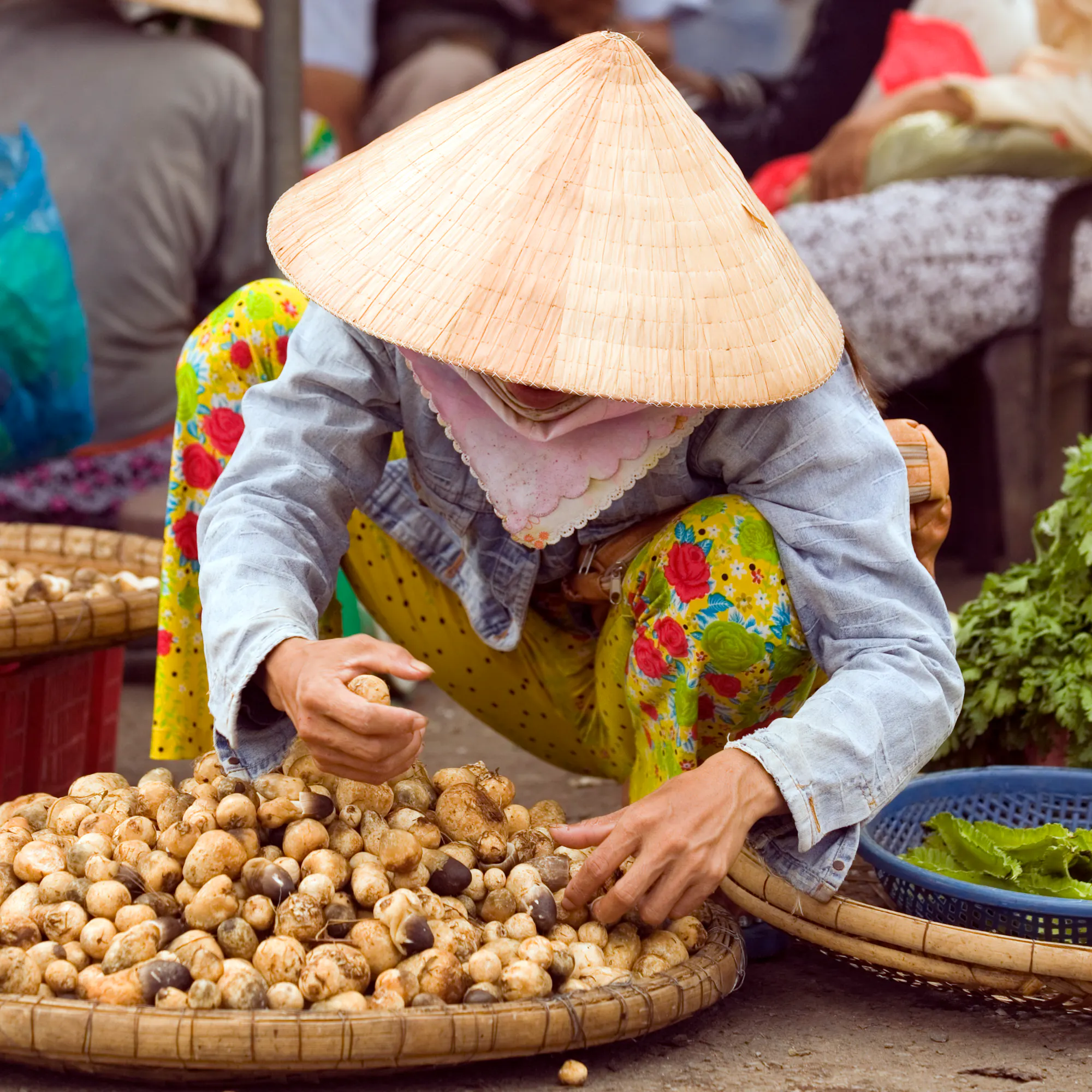 A woman in a conical hat sells vegetables from baskets spread on the ground at a street market.