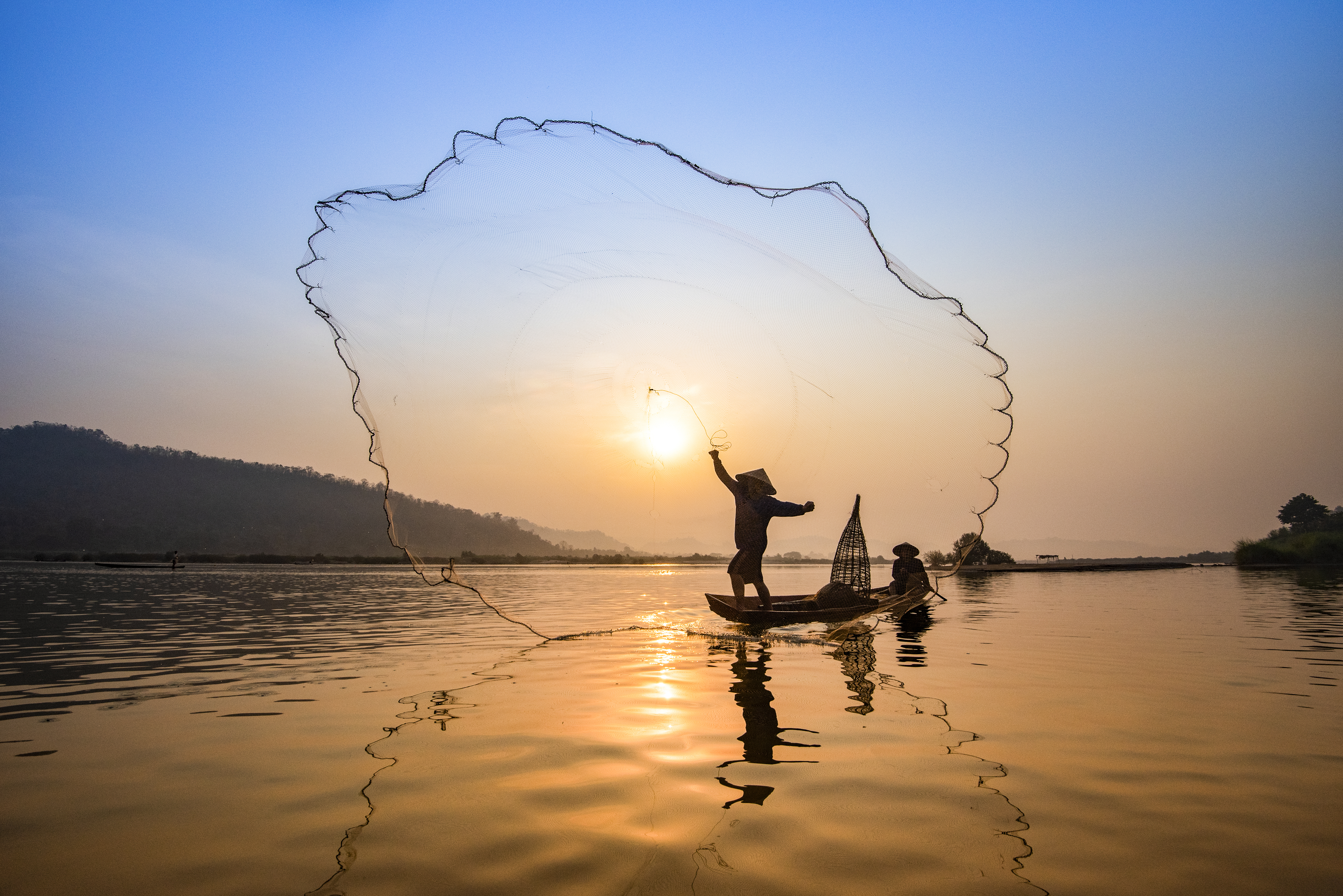 The silhouette of a fisherman casting a net from a boat is seen against a blazing Mekong sunset.