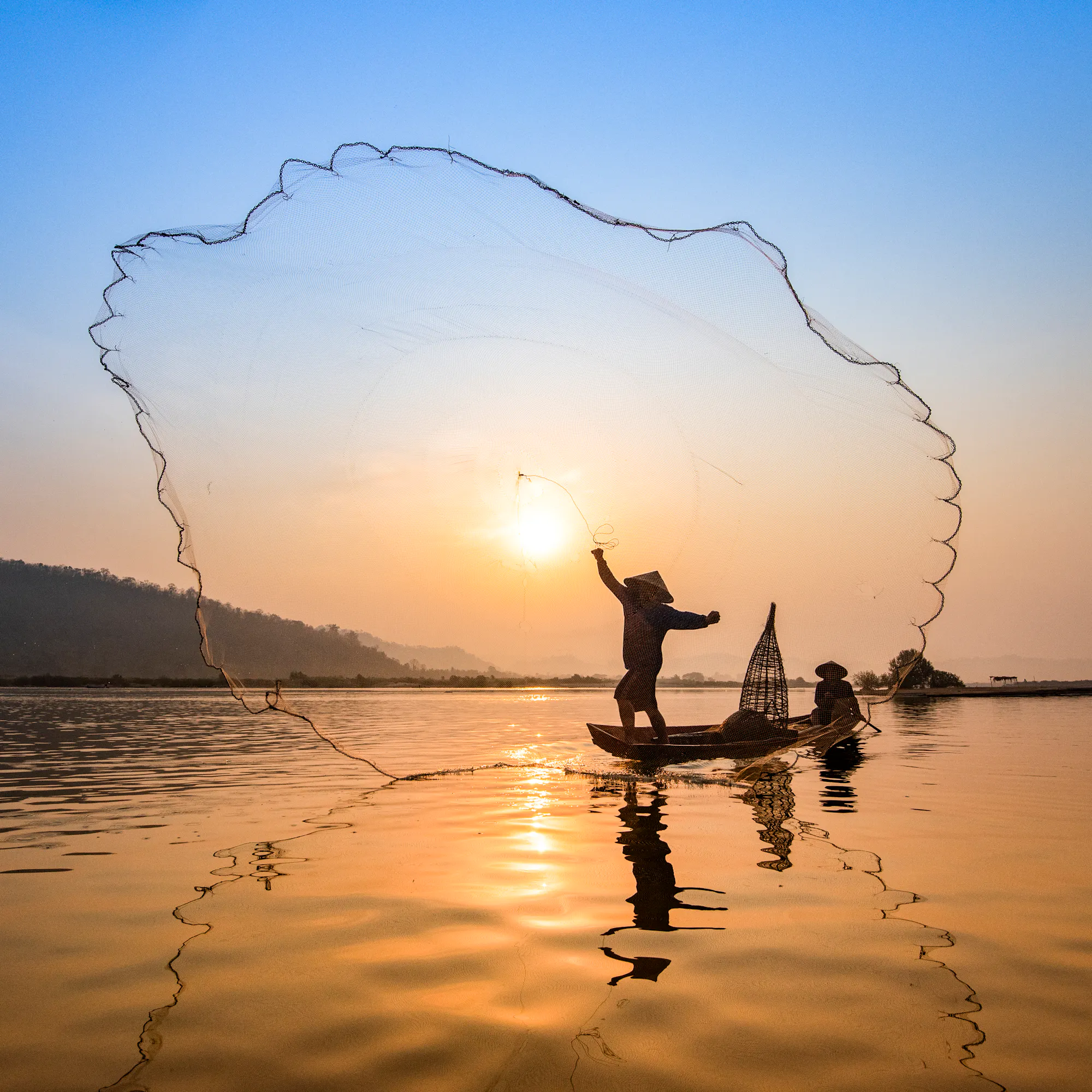 The silhouette of a fisherman casting a net from a boat is seen against a blazing Mekong sunset.