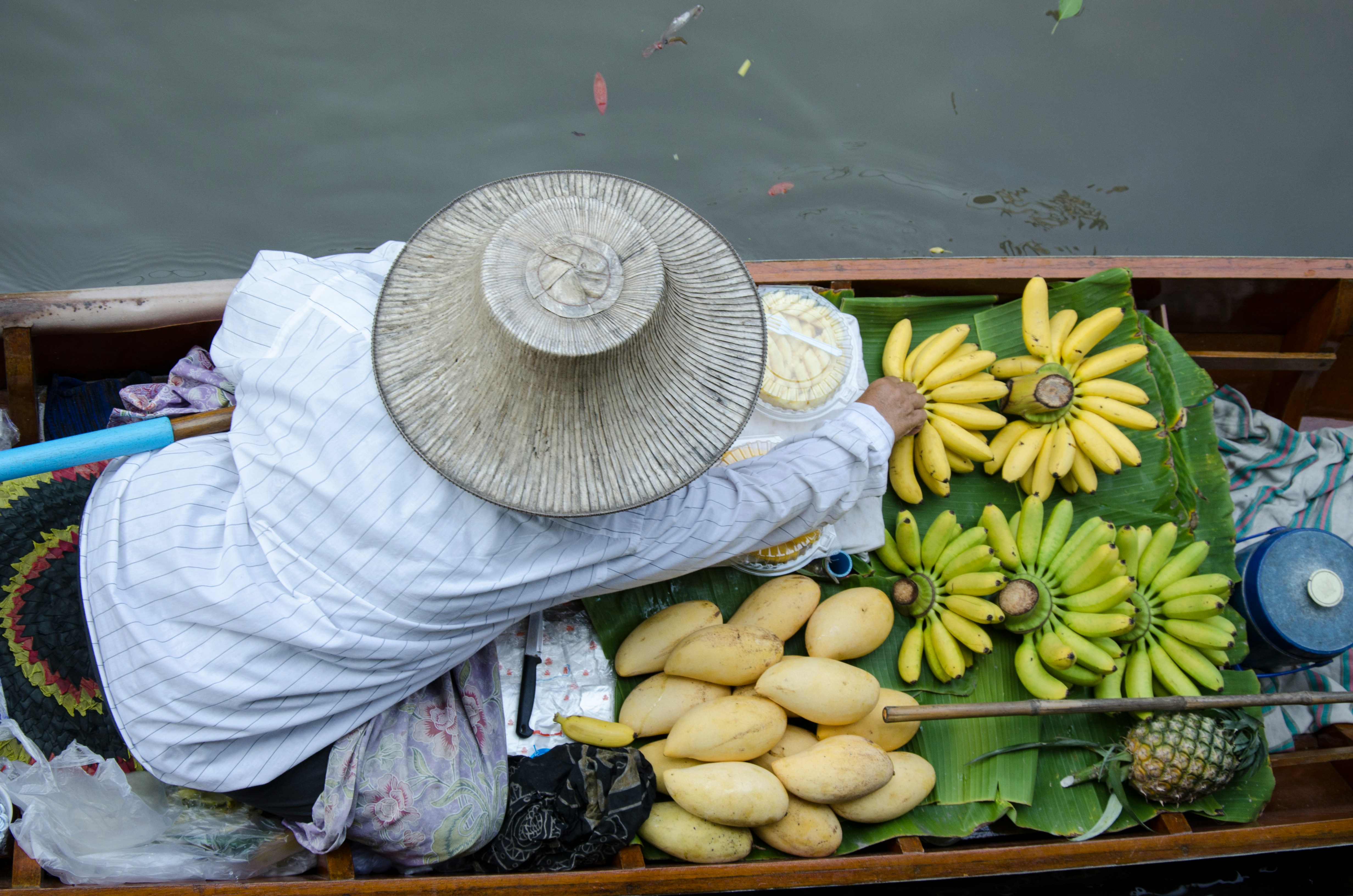 A woman in a wide-brimmed hat sells sunflowers and produce from a wooden cart on a Thai street.