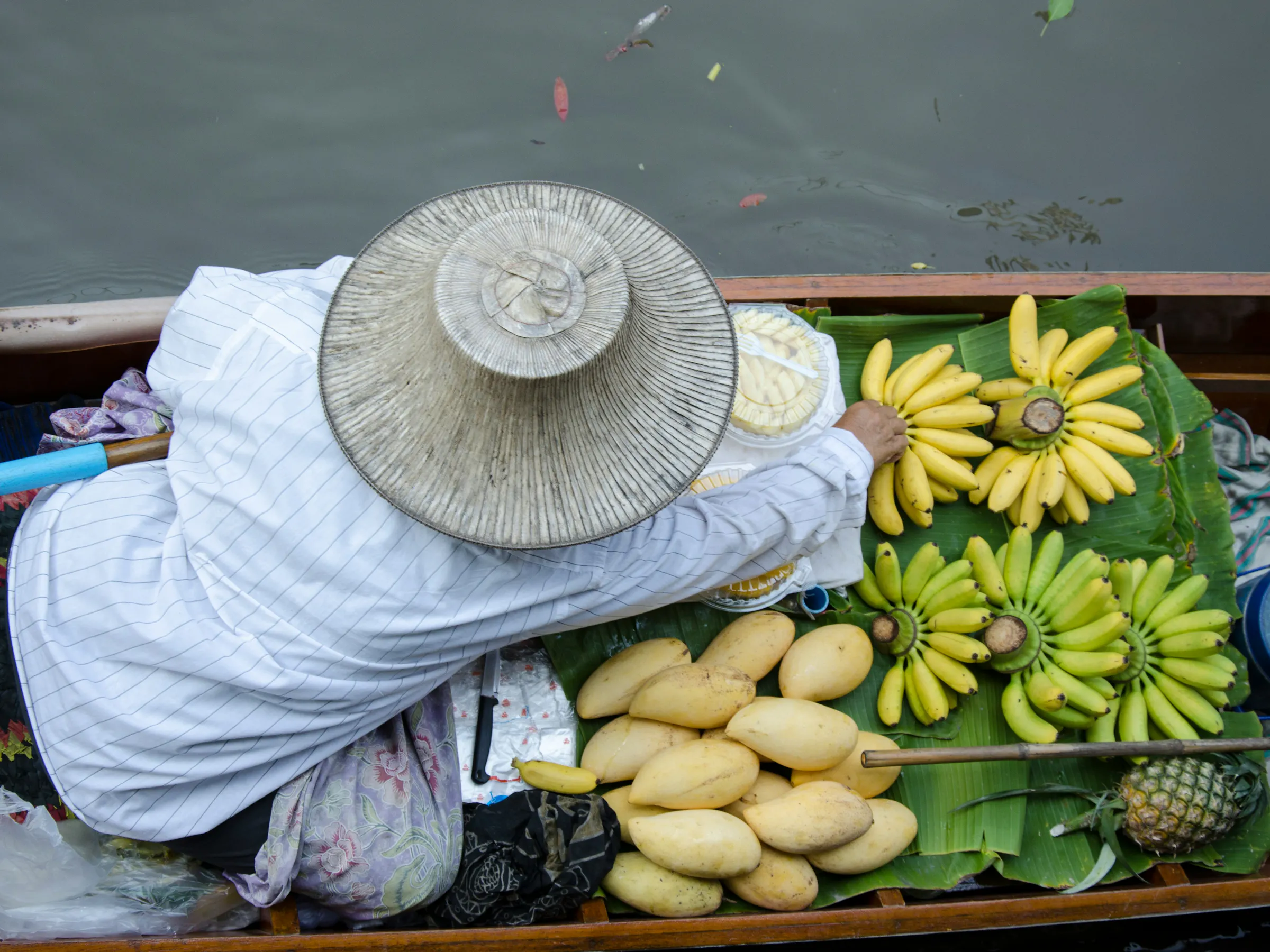 A woman in a wide-brimmed hat sells sunflowers and produce from a wooden cart on a Thai street.