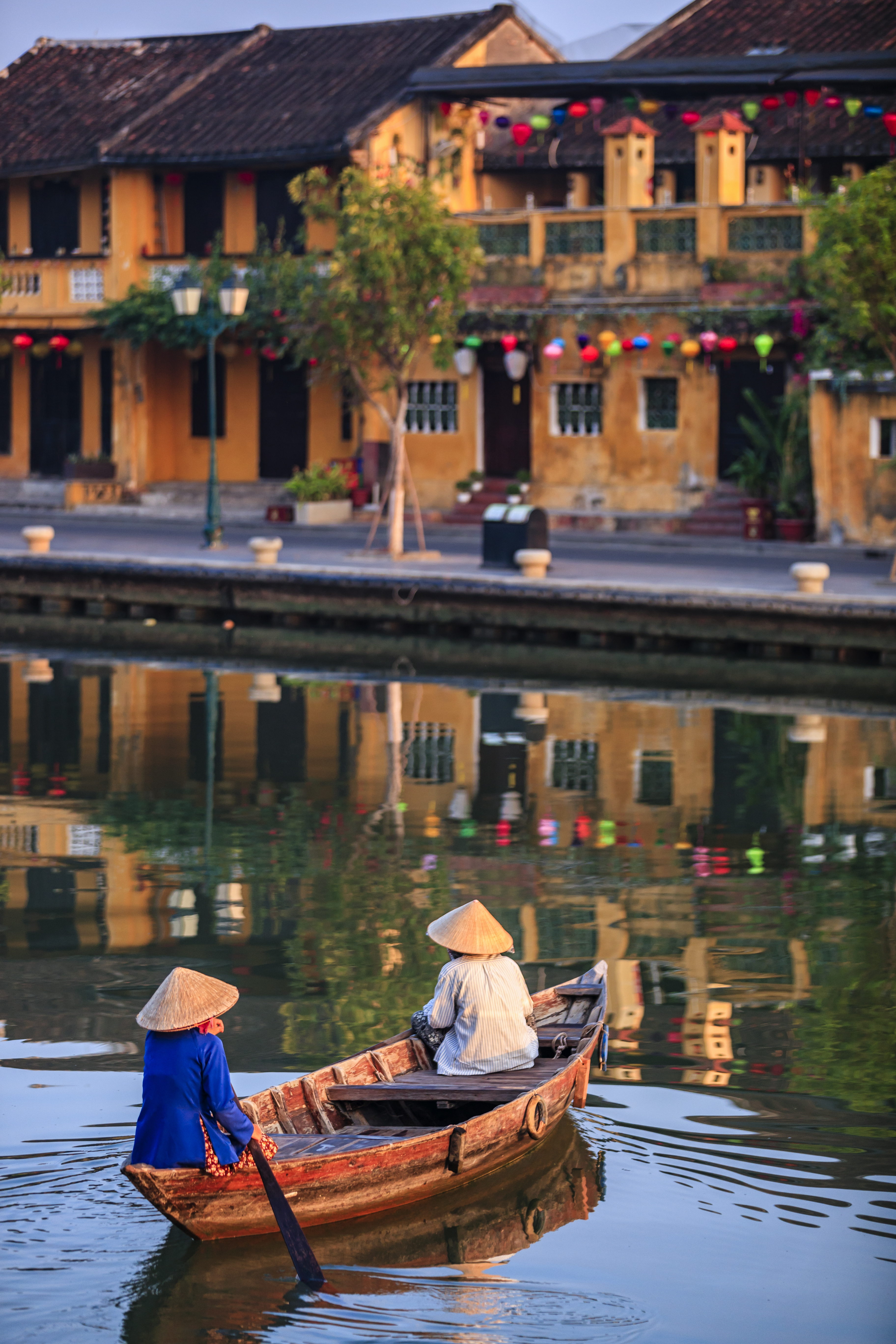 Two women in conical hats row a wooden boat through a reed-lined canal in Hoi An's old town.
