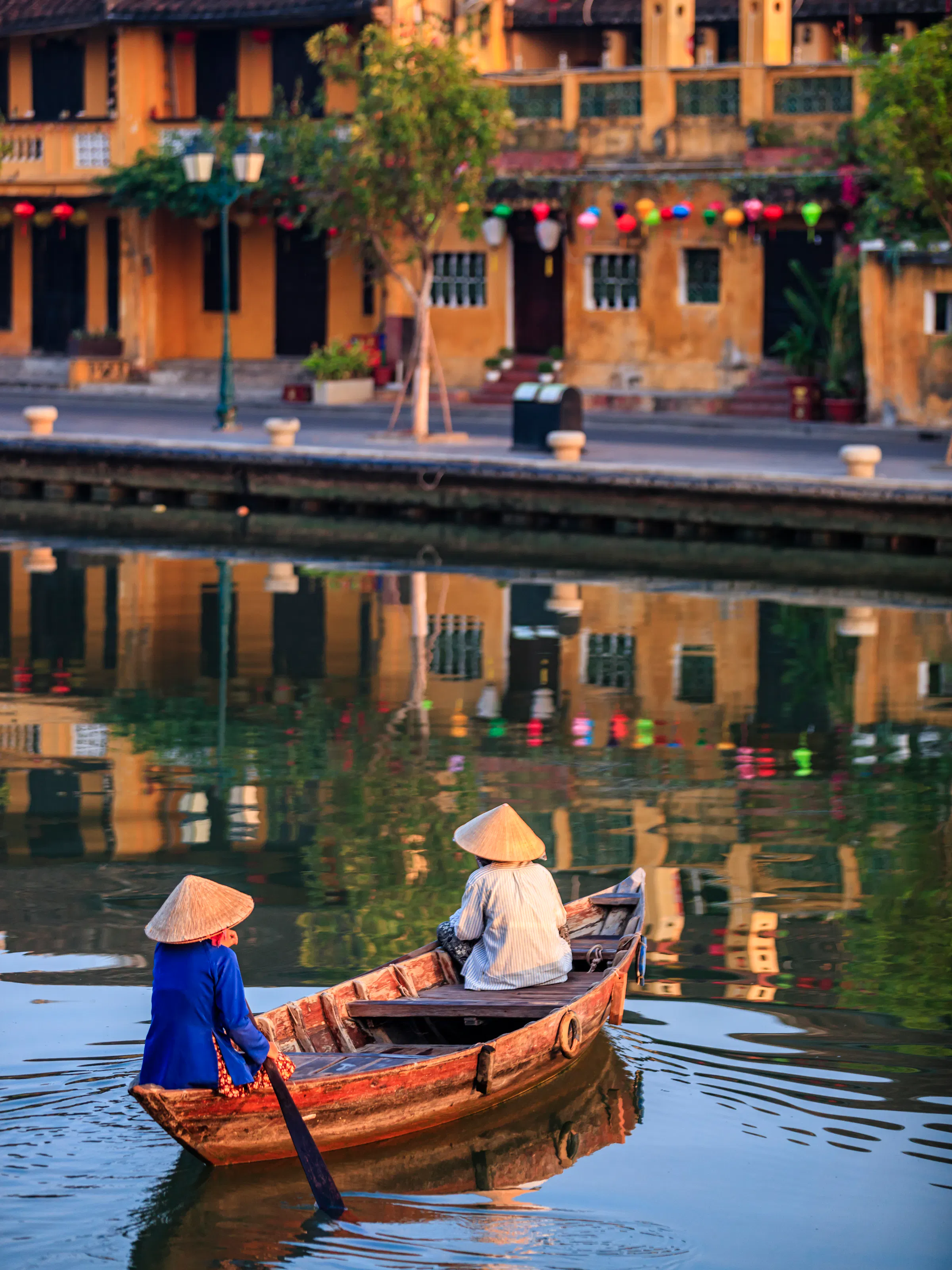 Two women in conical hats row a wooden boat through a reed-lined canal in Hoi An's old town.
