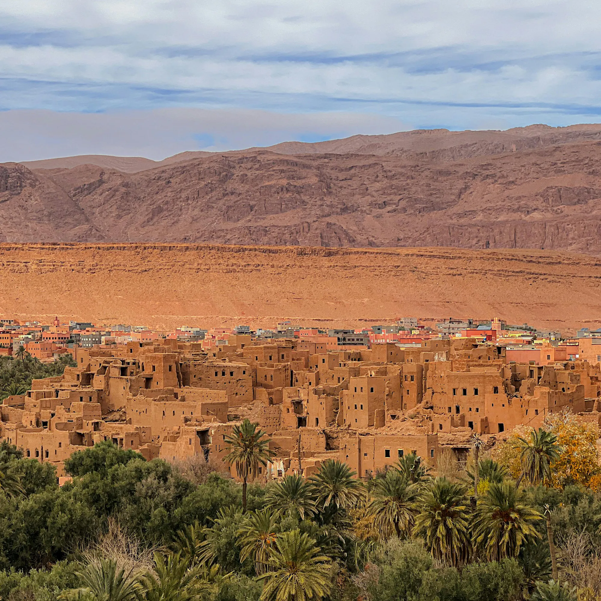 A mud-brick kasbah rises above a sparse desert landscape with low hills and dry scrub in Ouarzazate.
