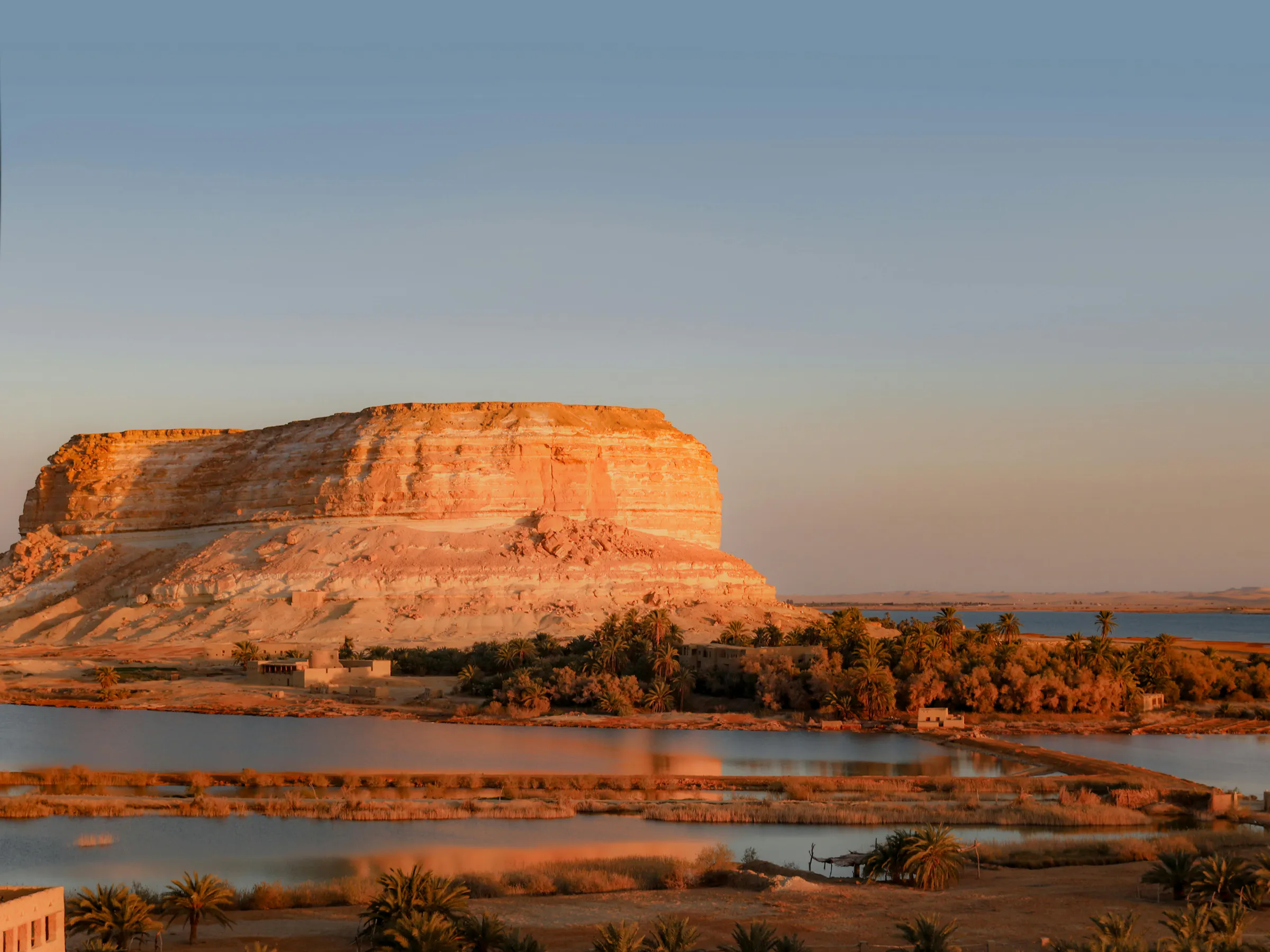 A flat desert mesa rises above a shallow lake and arid plain in the Siwa Oasis region of Egypt.