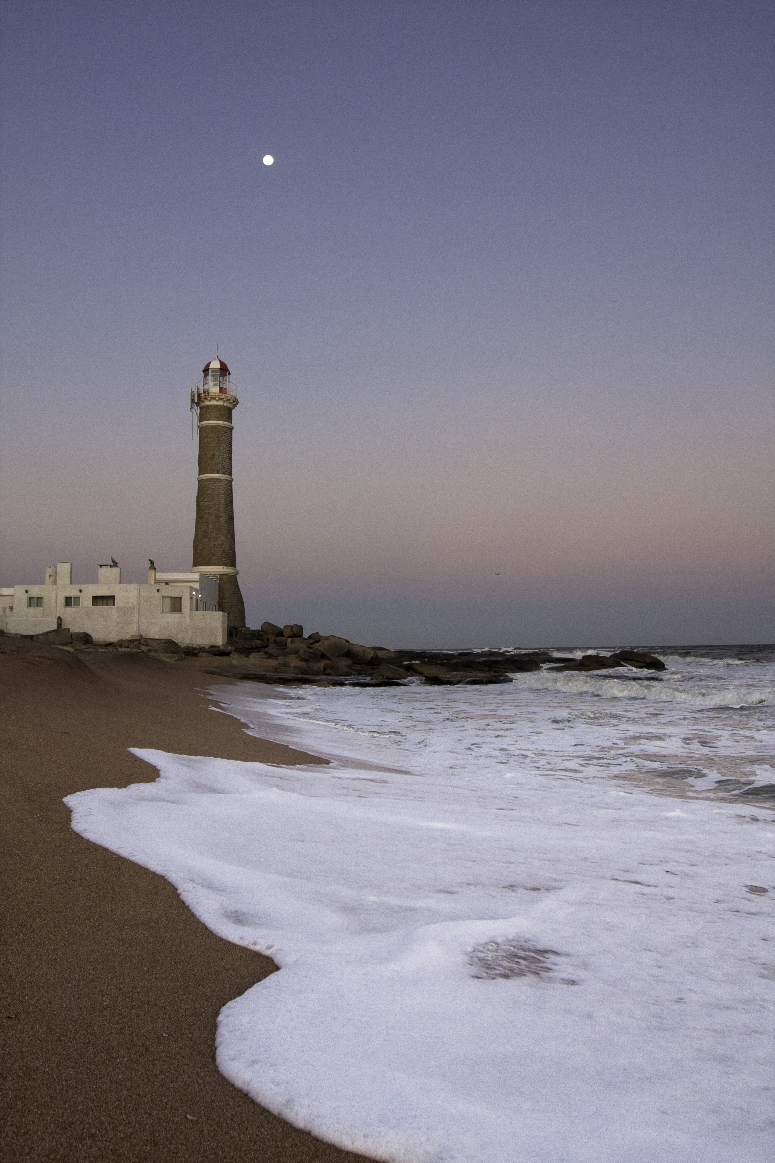 A solitary lighthouse stands on a sandy beach with gentle surf and a hazy horizon in Punta del Este.