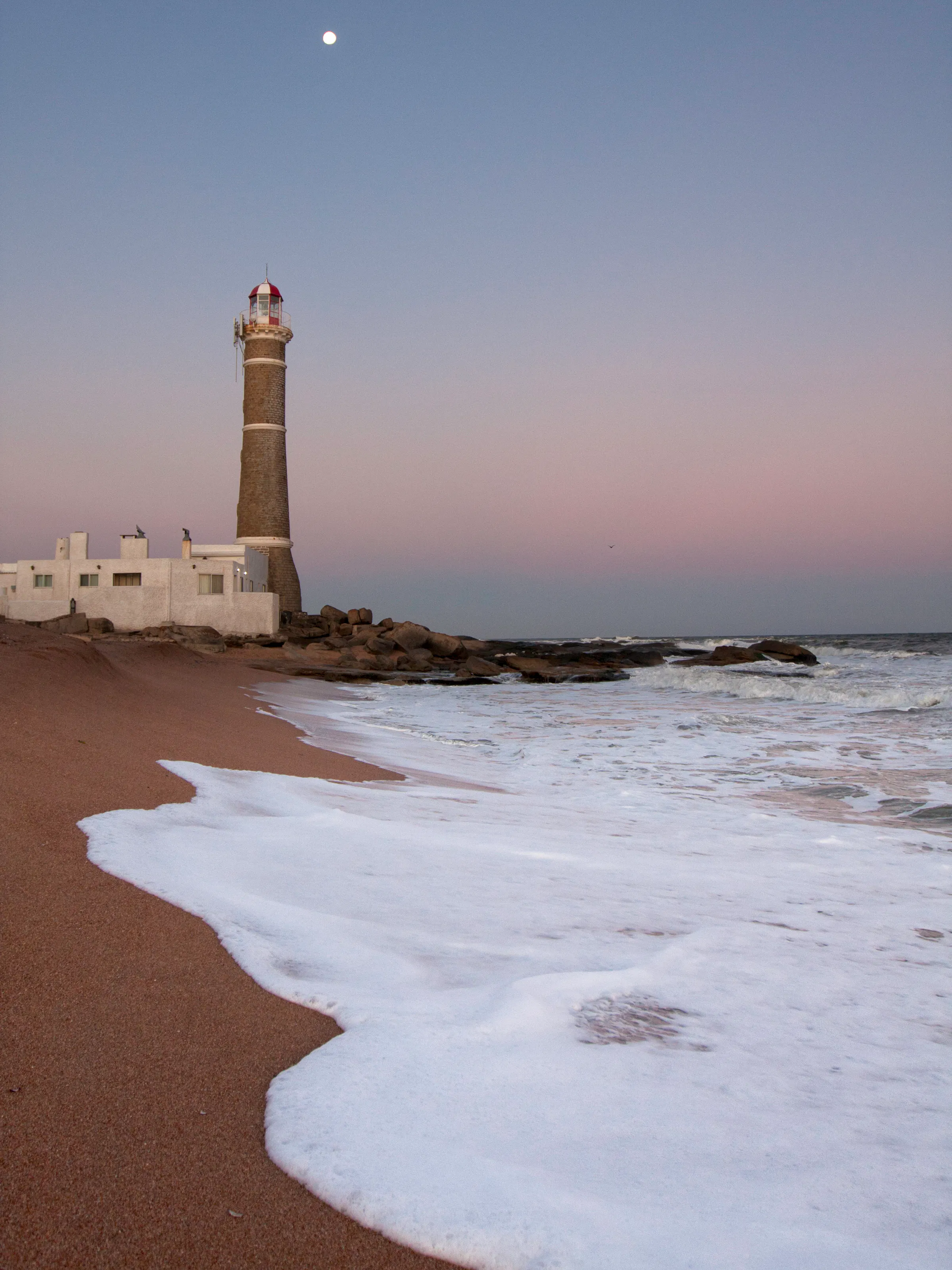 A solitary lighthouse stands on a sandy beach with gentle surf and a hazy horizon in Punta del Este.