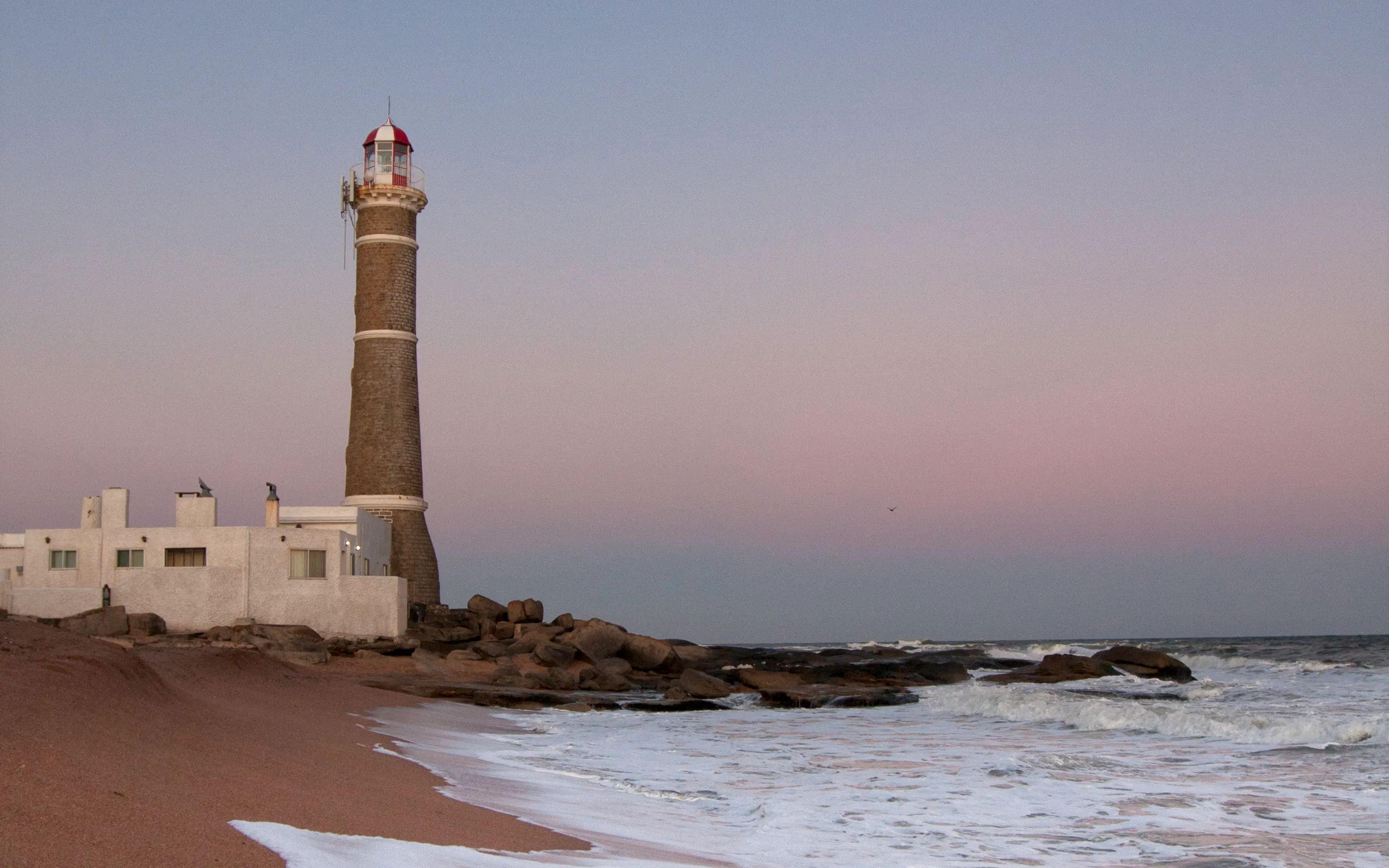 A solitary lighthouse stands on a sandy beach with gentle surf and a hazy horizon in Punta del Este.