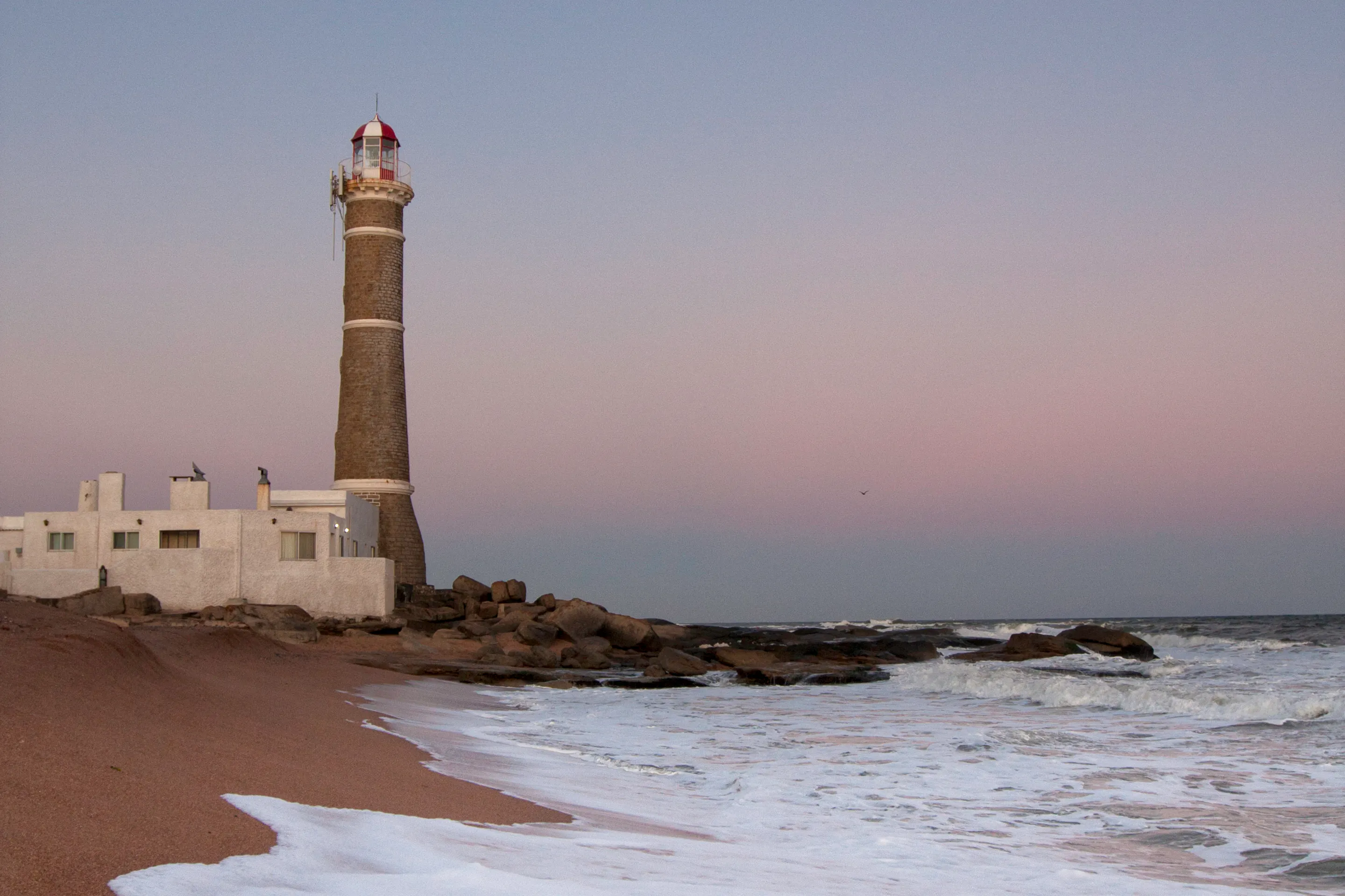 A solitary lighthouse stands on a sandy beach with gentle surf and a hazy horizon in Punta del Este.