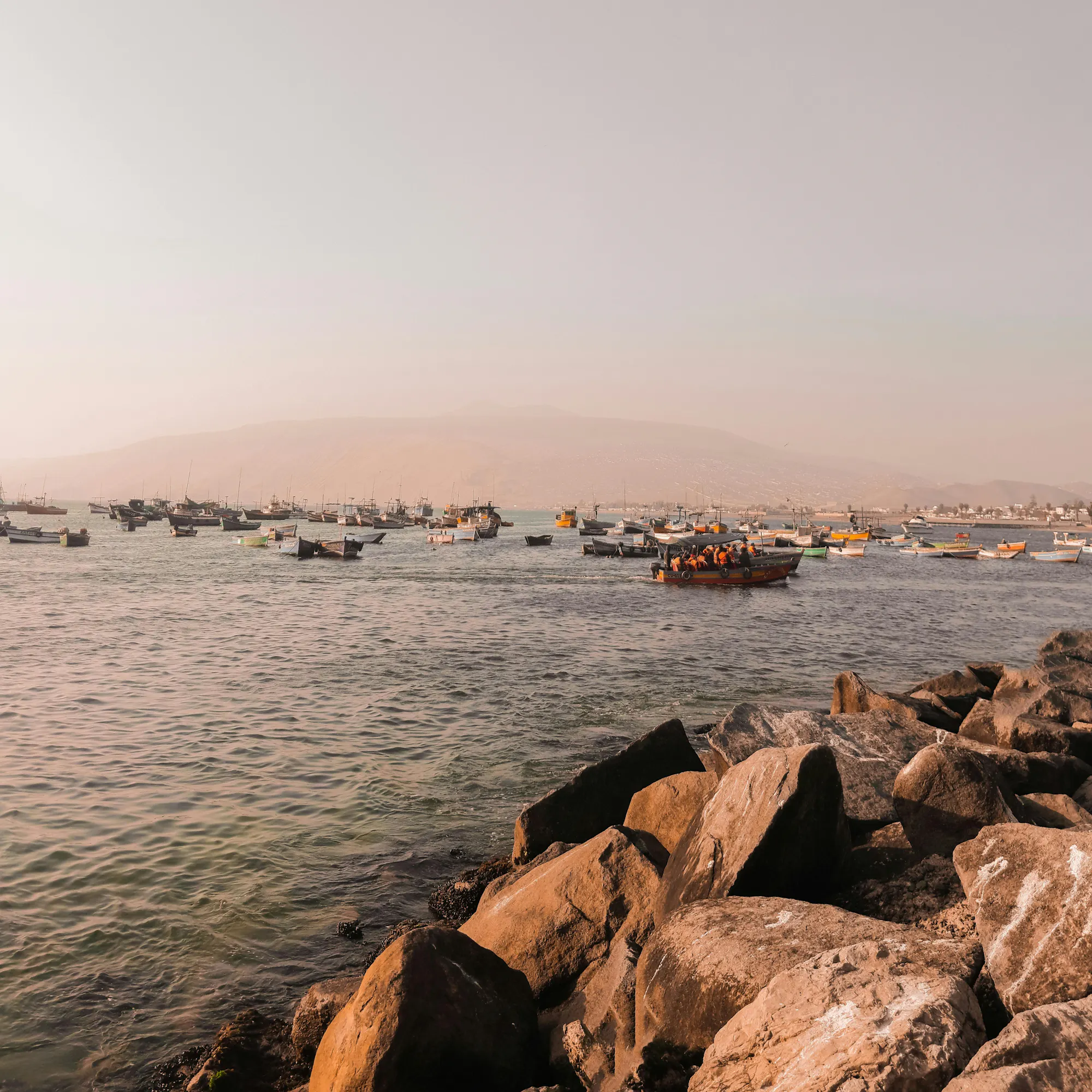 Rocky boulders line the Pacific shoreline at Lima with a hazy sky and calm grey sea beyond.