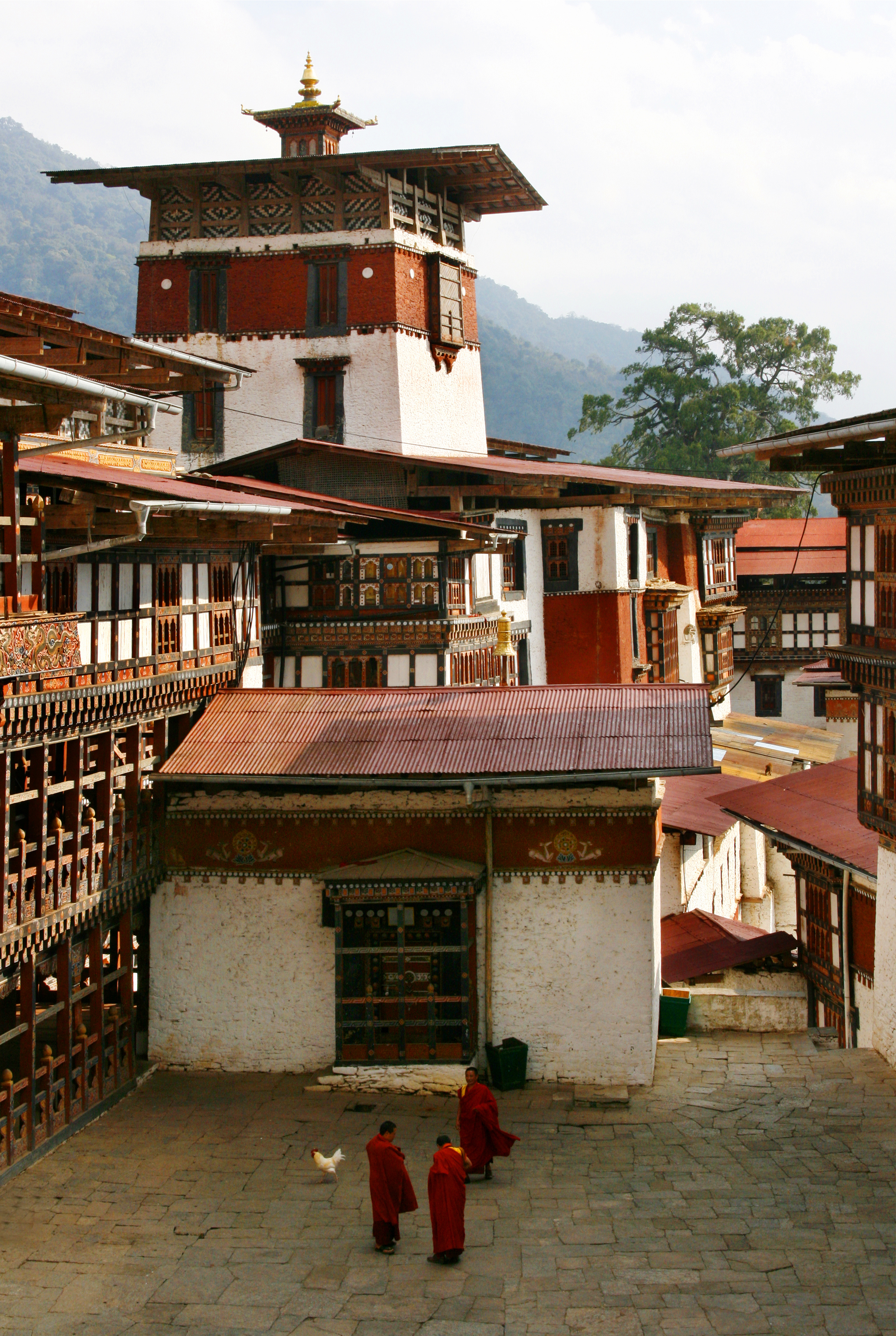A traditional Bhutanese dzong with white walls and a red roof band sits amid mountain scenery.
