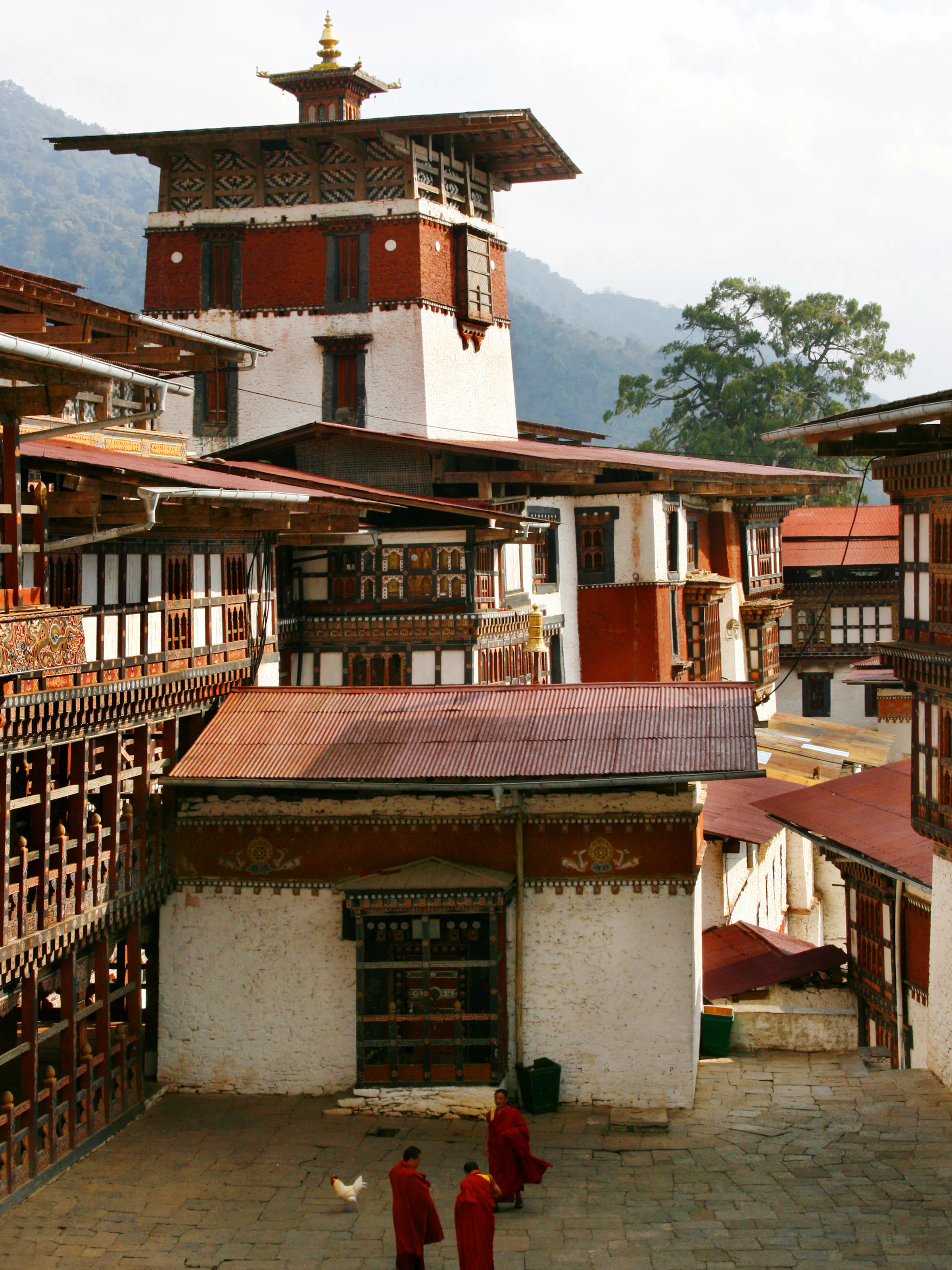 A traditional Bhutanese dzong with white walls and a red roof band sits amid mountain scenery.