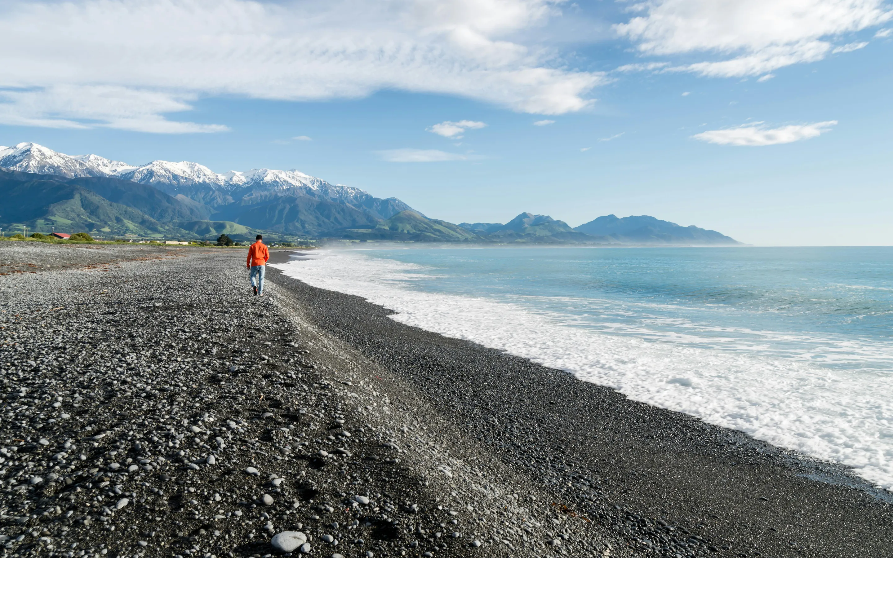 A lone figure stands on a dark pebble beach looking toward distant mountains under a pale sky.