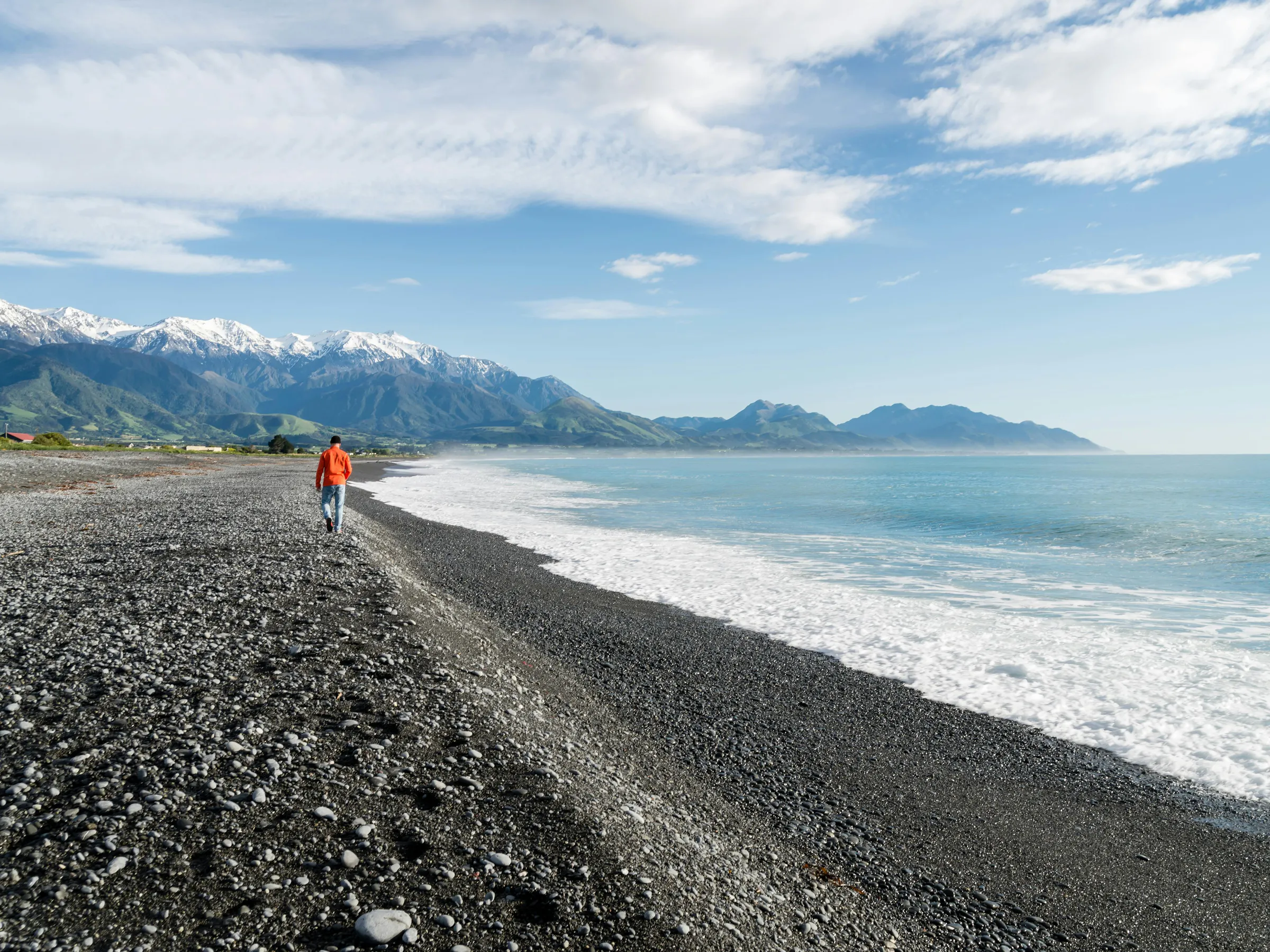 A lone figure stands on a dark pebble beach looking toward distant mountains under a pale sky.